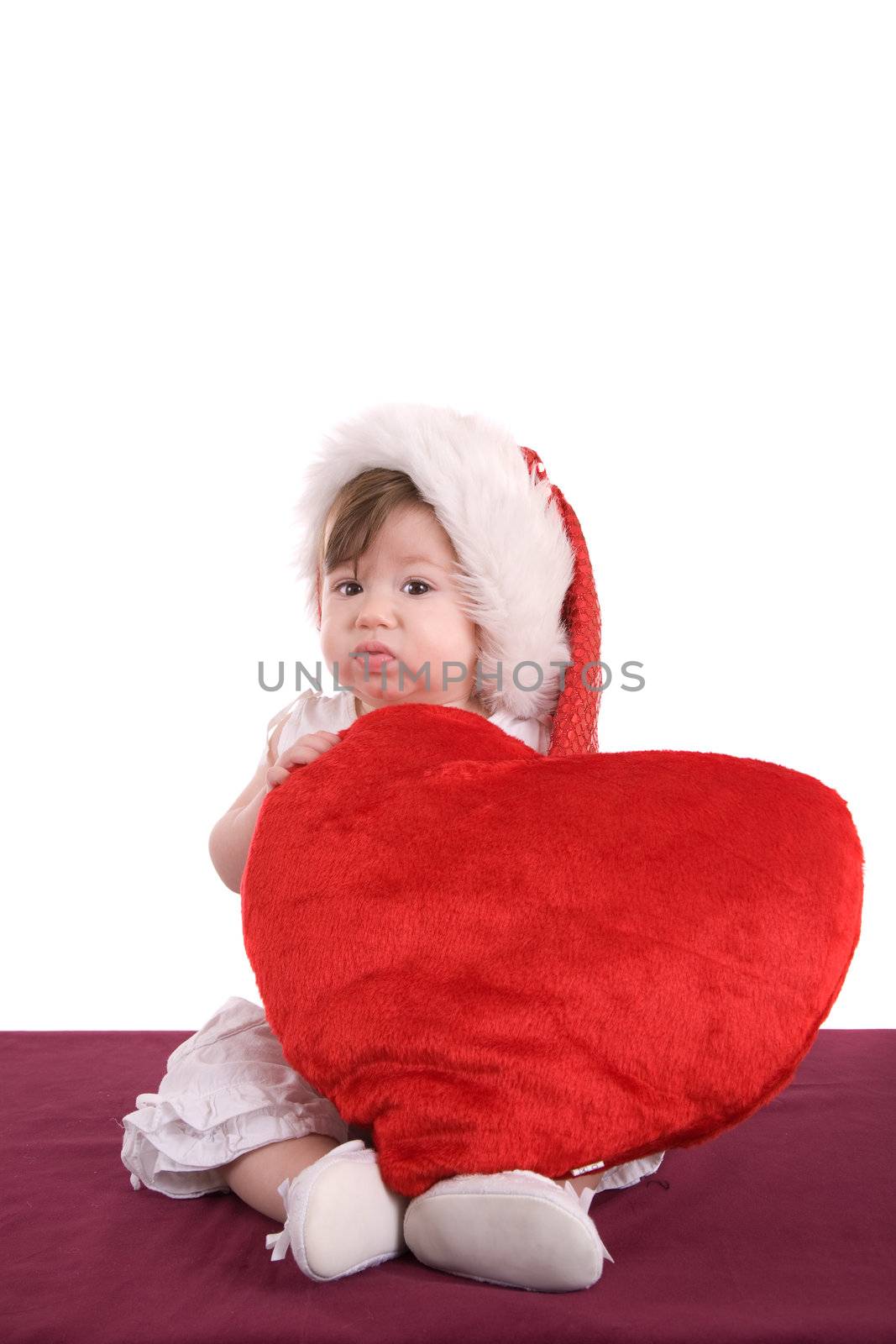 Cute baby girl sitting with a christmas hat and big red heart not looking very happy