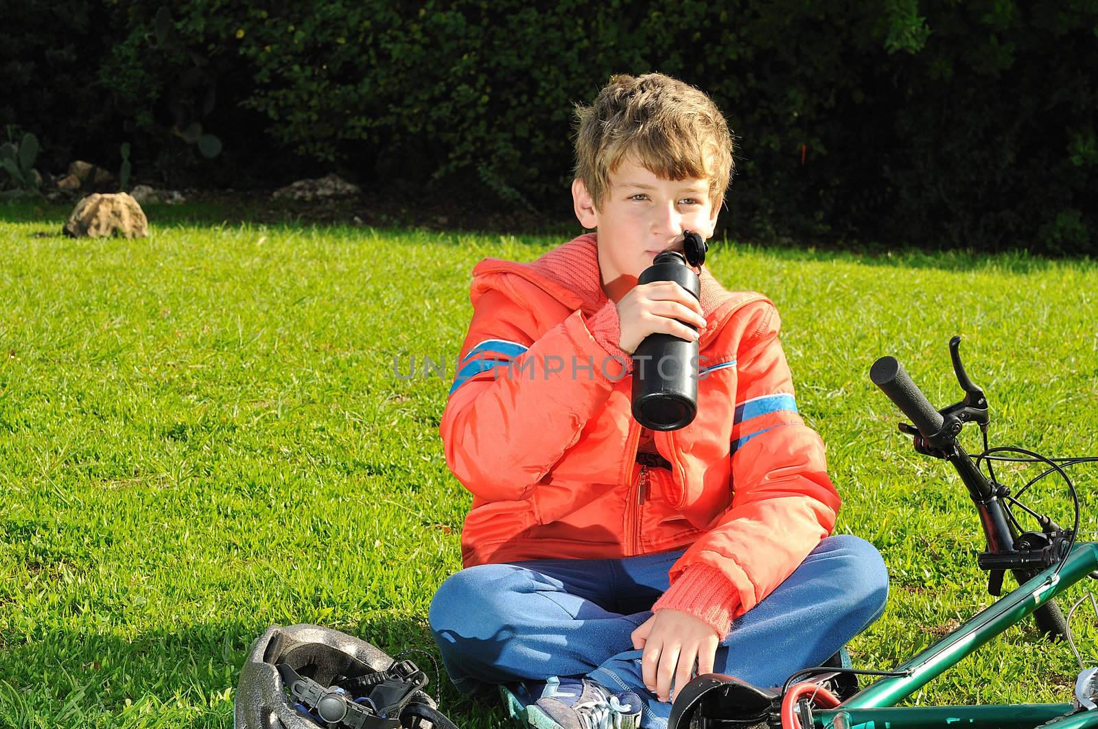 
The boy sitting on the grass in a park near the bike and drinking water from a bottle