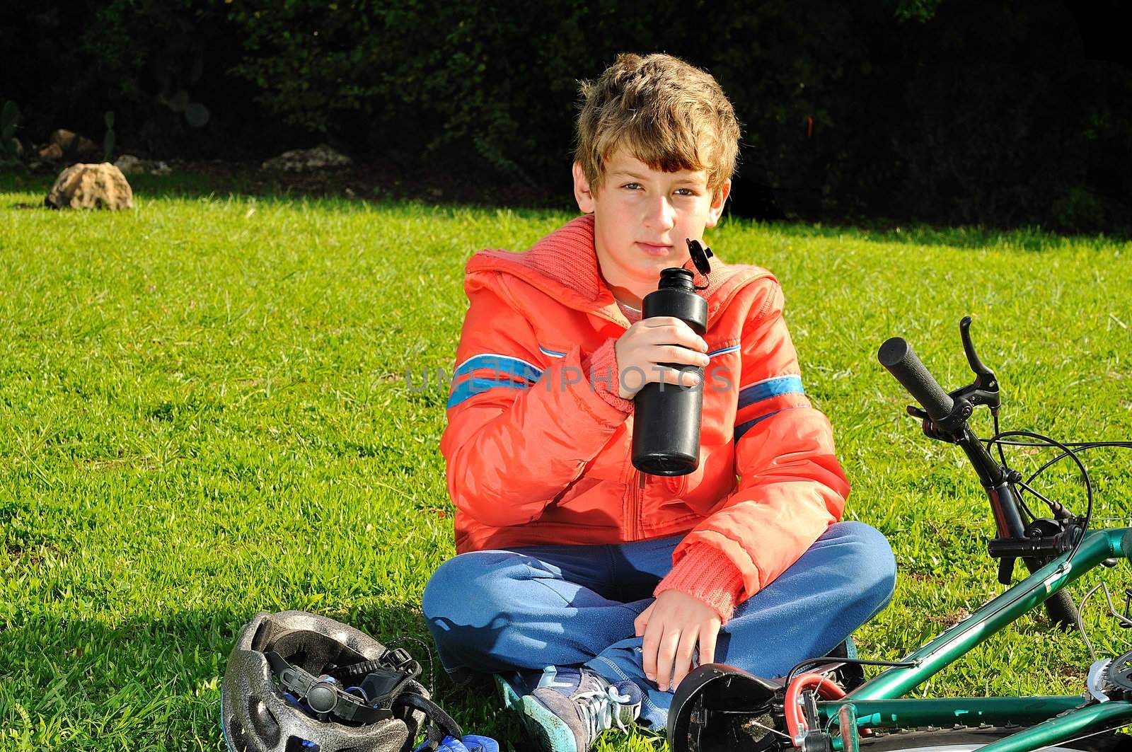 
The boy sitting on the grass in a park near the bike and drinking water from a bottle