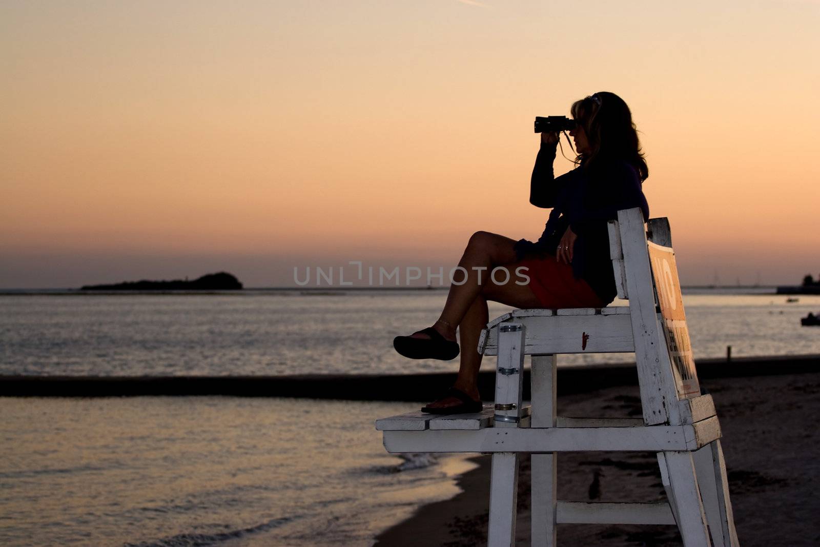 A silhouette of a woman looking with binoculars at the beach while sitting on a lifeguard chair.