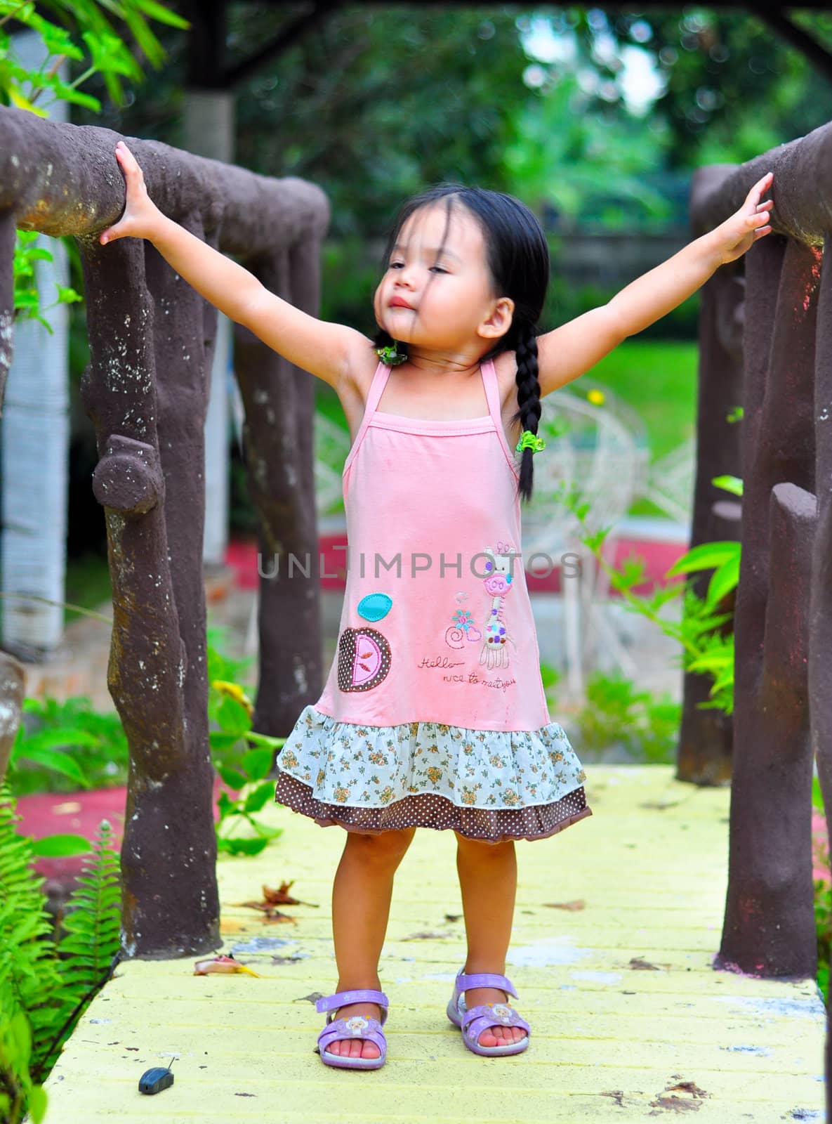 A cute little girl wearing a pink dress, standing with arms wide open on a bridge.