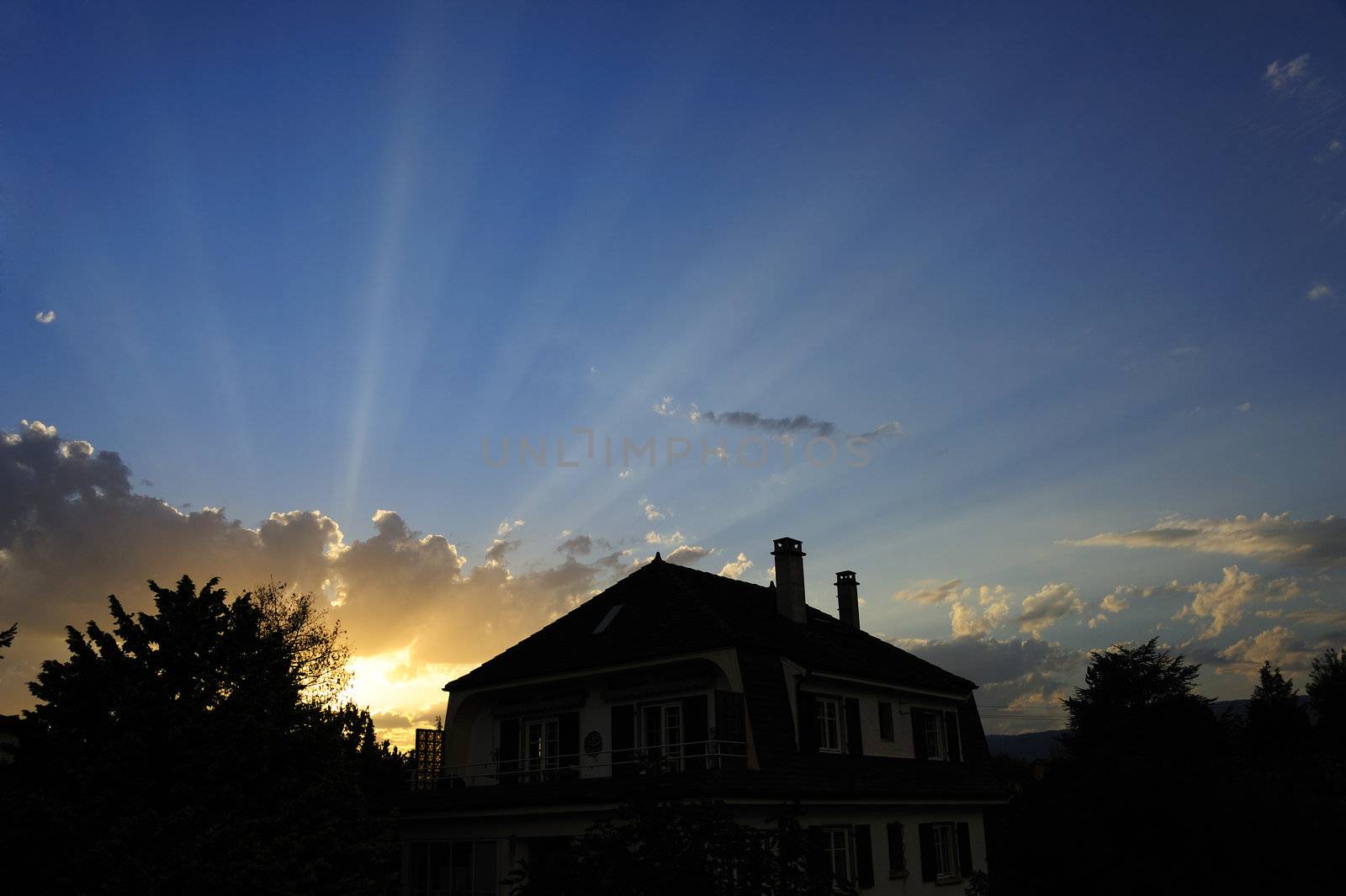 The sun rising over (or setting behind) the silhouette of a house. Crepuscular rays streaming out from behind the clouds. Space for text in the sky.