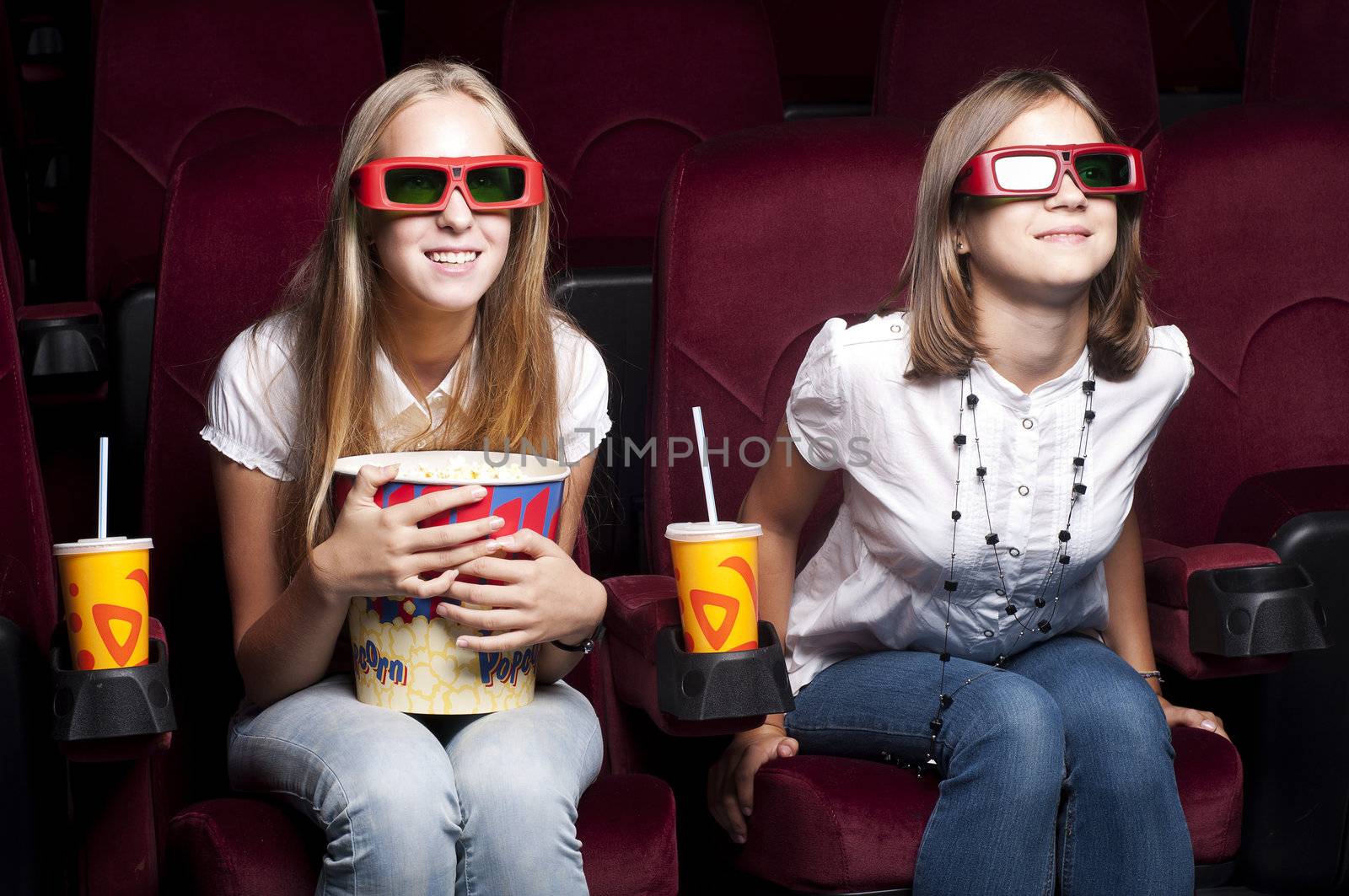 two girls look three-dimensional cinema, sitting in the glasses, eat popcorn, drink drink