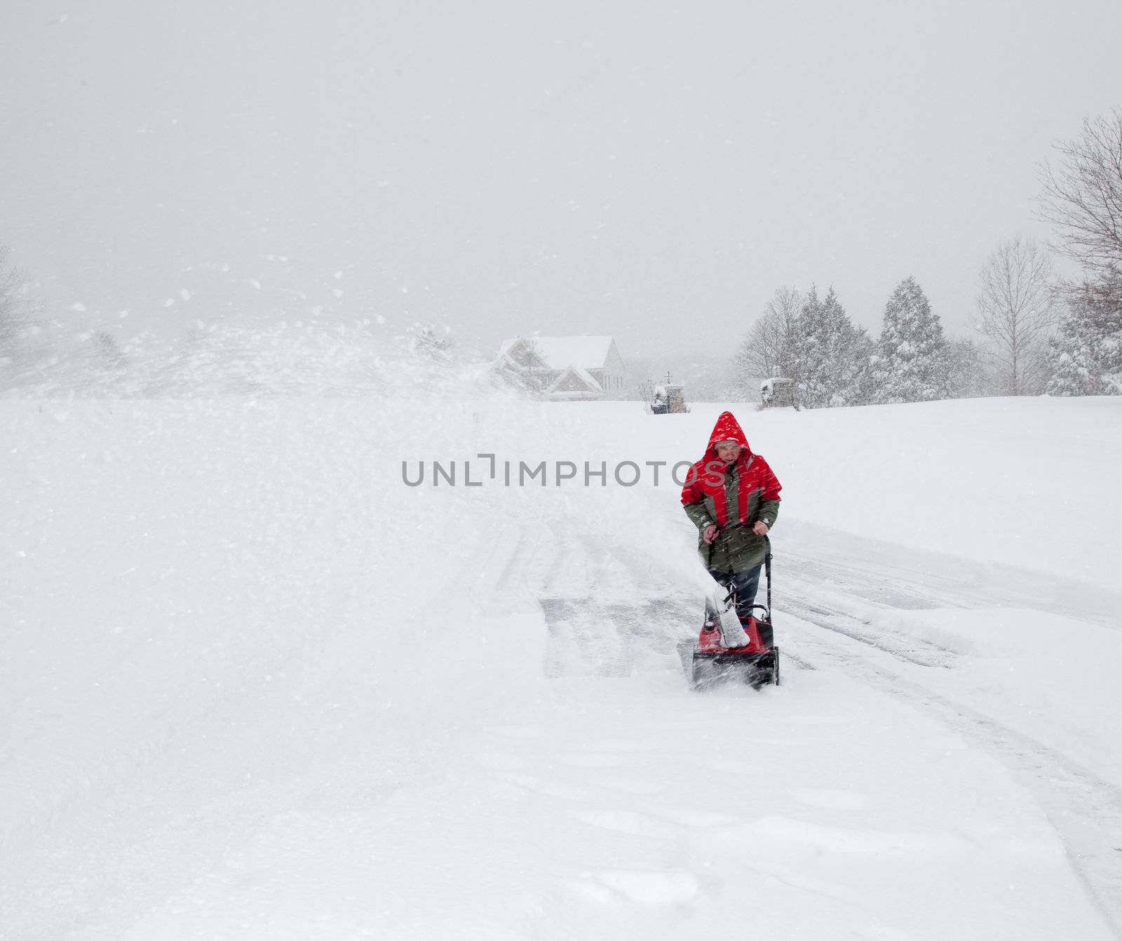 Senior man in red coat using a snow blower during a blizzard on home drive