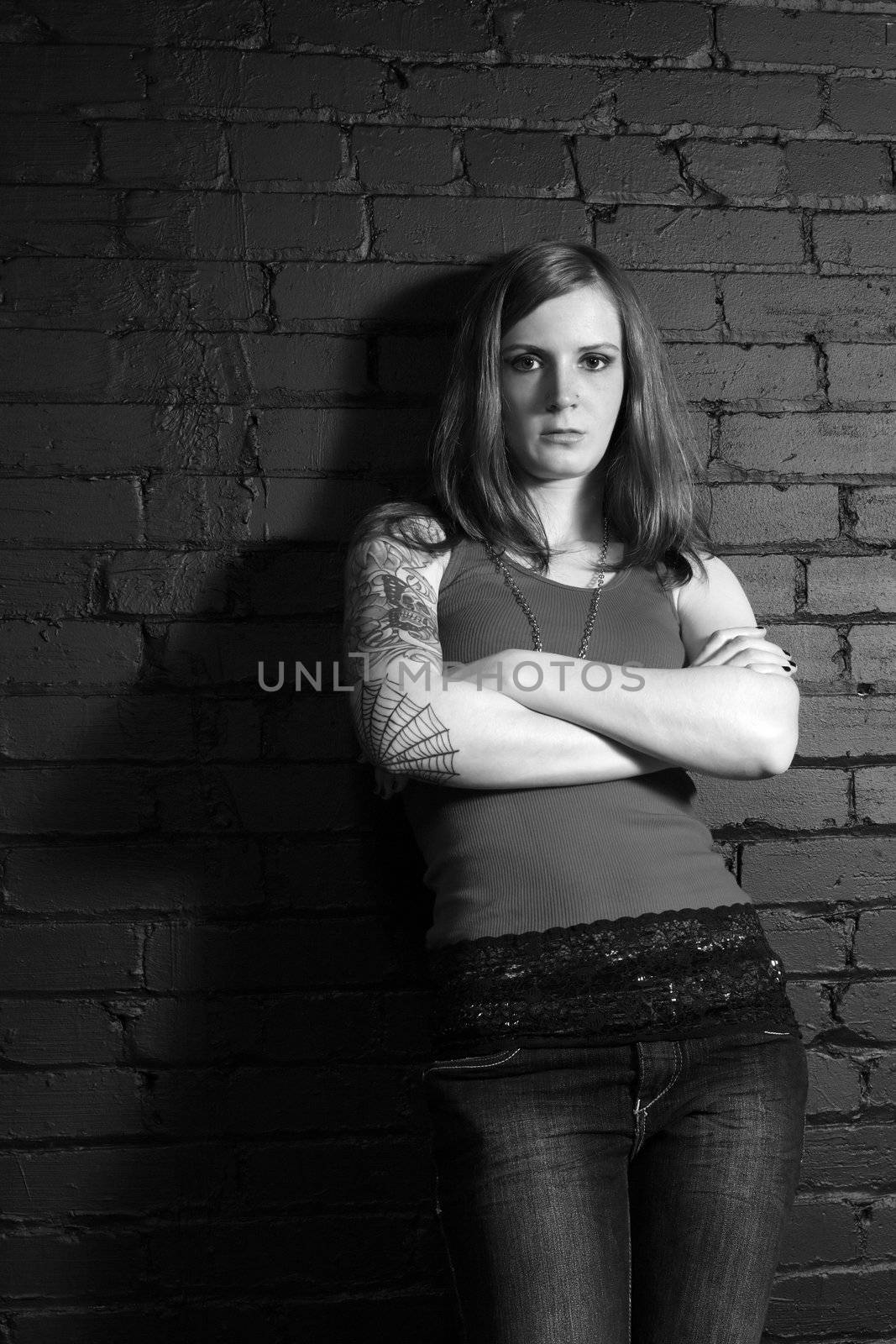 A young female with full arm tattoo leaning up against a black brick wall.
