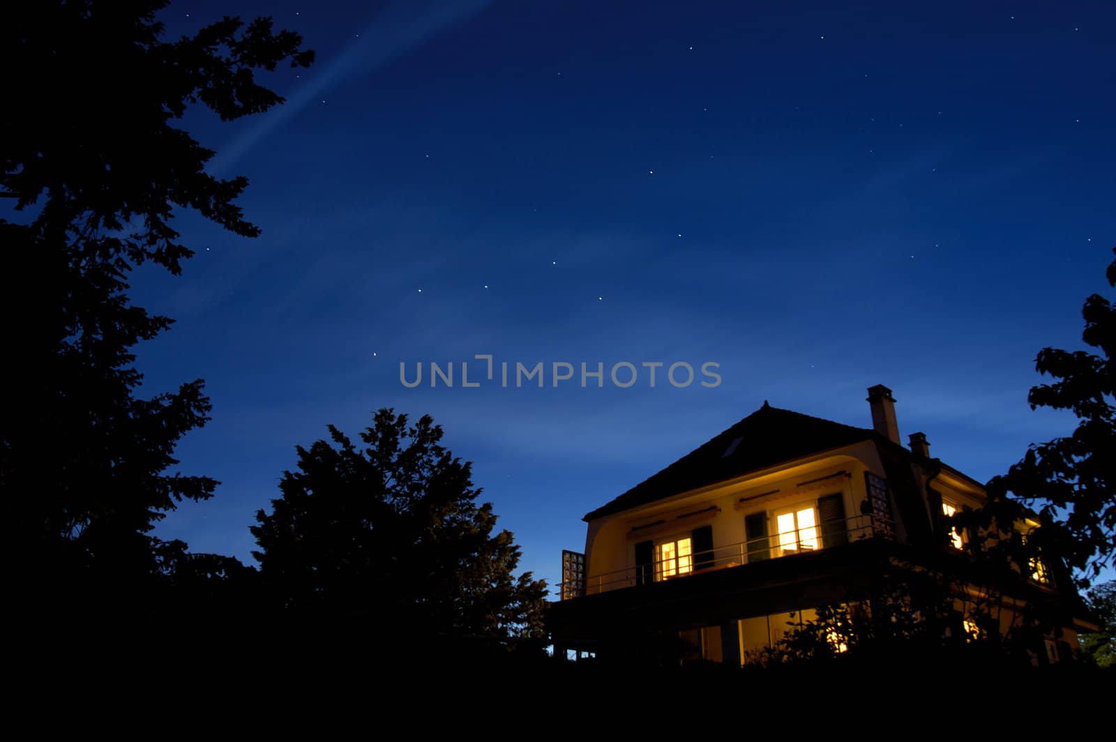 The constellation of The Plough hangs in the sky as the sun sets. Other stars come out whilst, beneath, the lights of a house glow warmly.