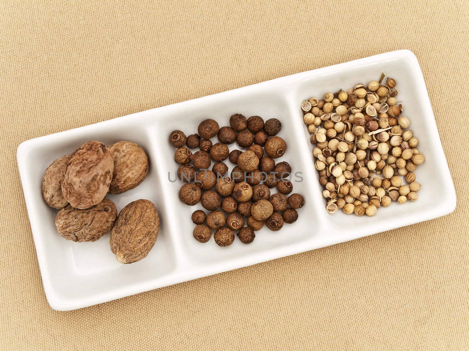 Coriander, black pepper seeds and nutmeg in flavouring plate at beige textile