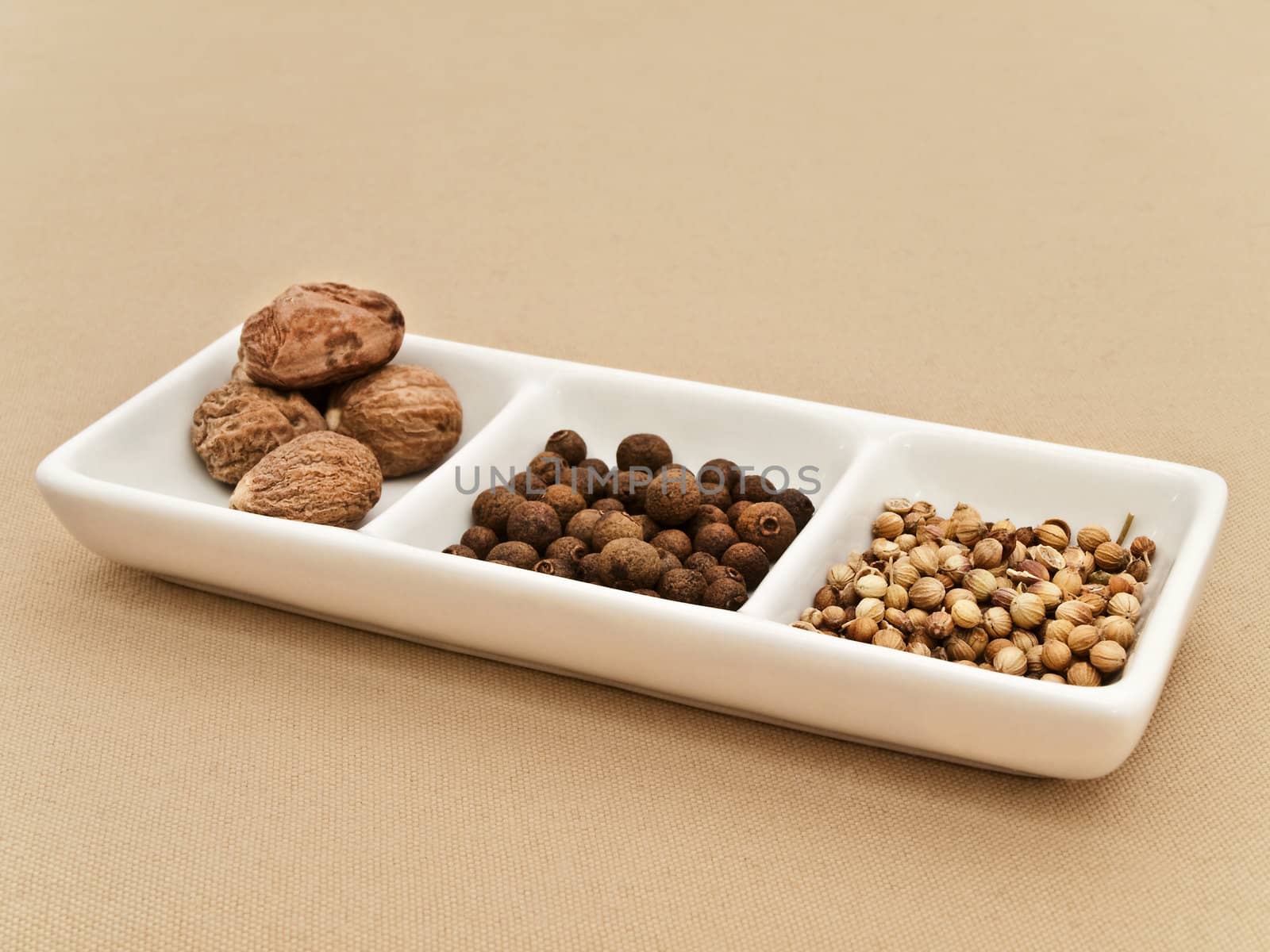 Coriander, black pepper seeds and nutmeg in flavouring plate at beige textile
