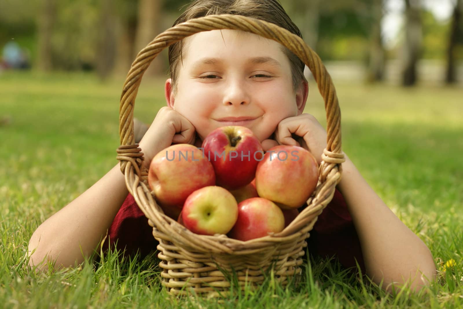 A child with a basketful of Pink Lady apples.  He is smiling and laying in the grass.