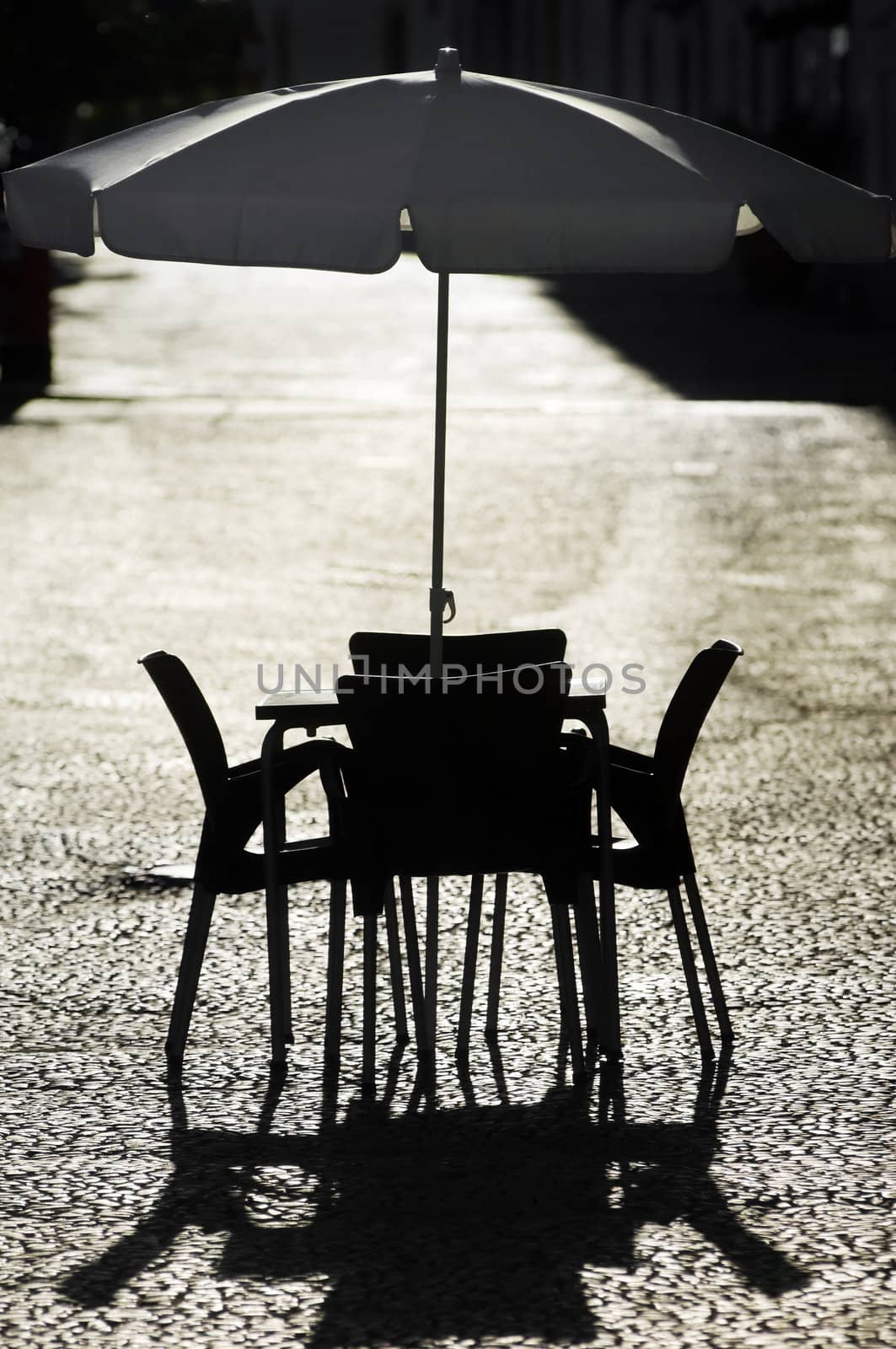A little empty coffee table with four chairs around and umbrella in the middle standing on the pavement of a sidewalk outdoors in atmospheric light. 