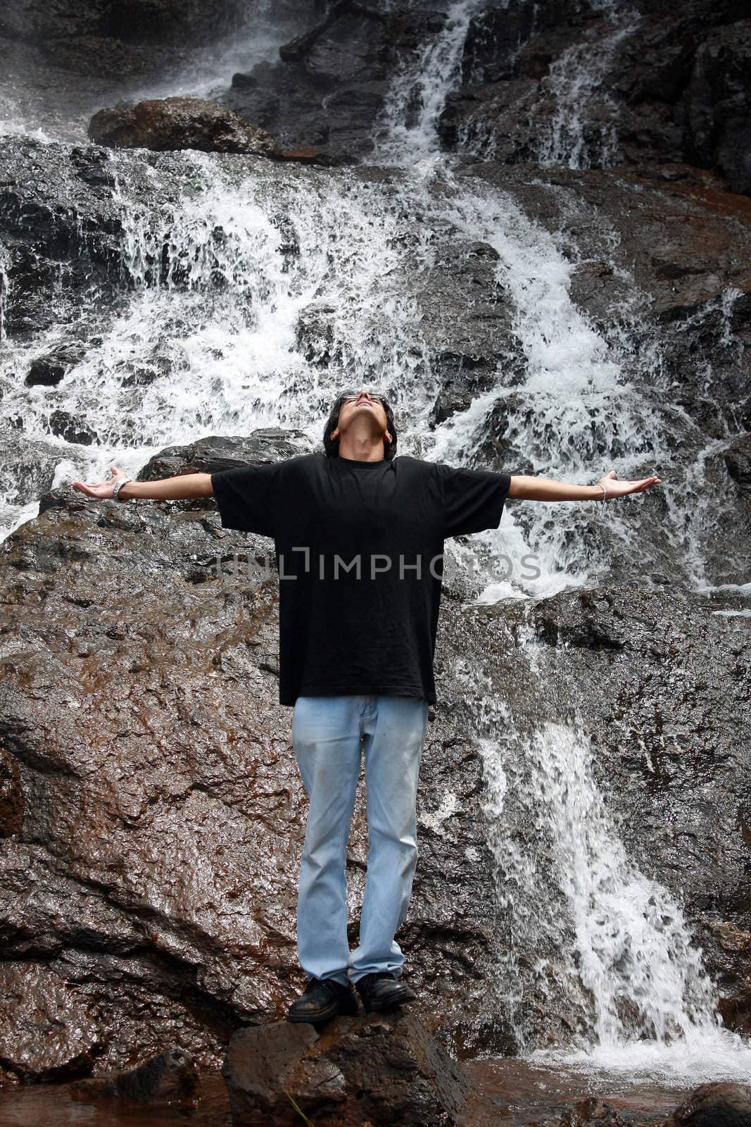 A young Indian guy standing with stretched arms, enjoying the coolness under a waterfall.