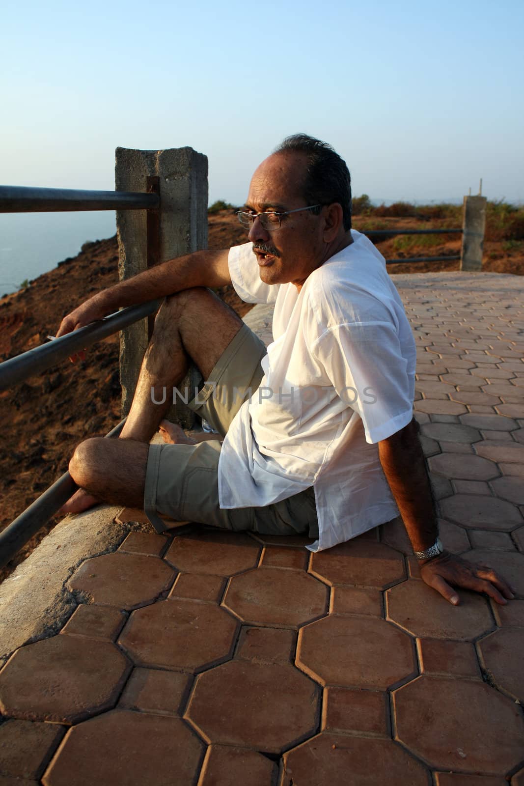 A relaxed senior Indian man sitting with his cigarette near a railing.