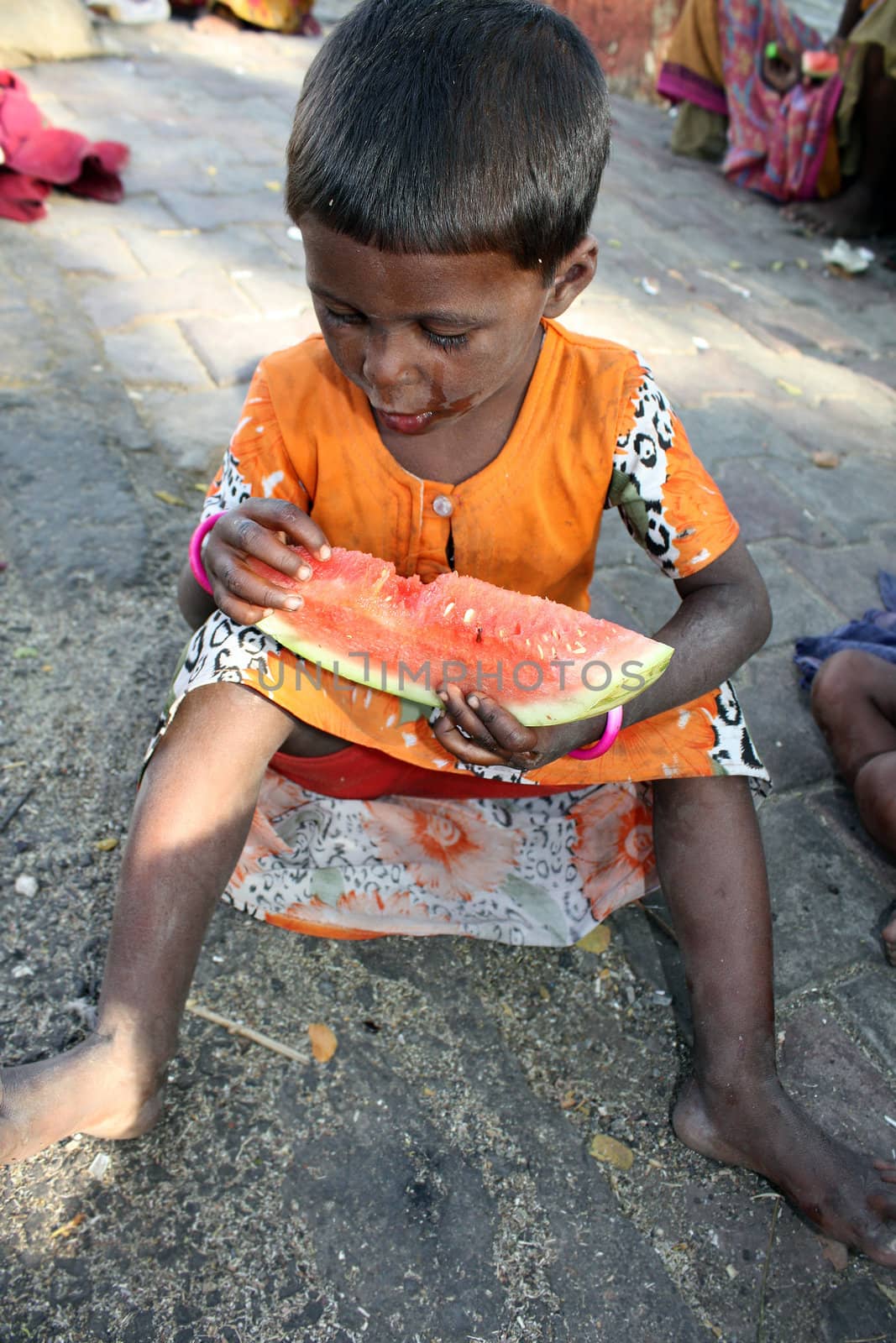 A hungry beggar girl from India eating a watermelon, sitting on a street.