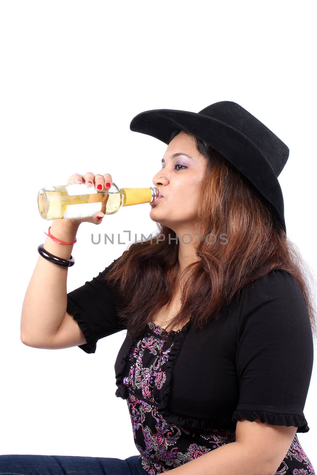 A young Indian woman in hat drinking a orange alcoholic beverage, on white studio background.