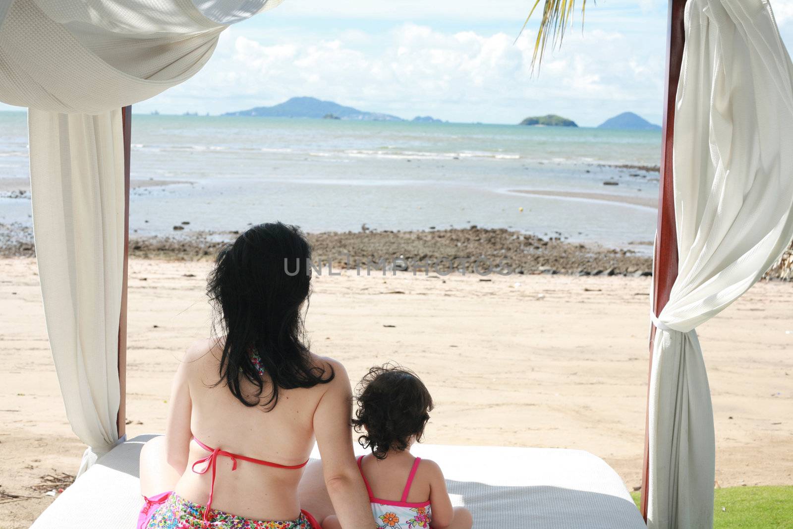 young lovely mother and her little daughter on tropical white bed close to the beach in the glow of the evening sun