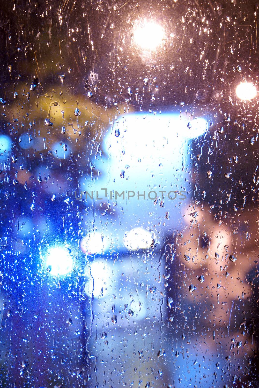 A full frame shot of a window looking outside towards the city lights while its raining, focus is on the rain drops.