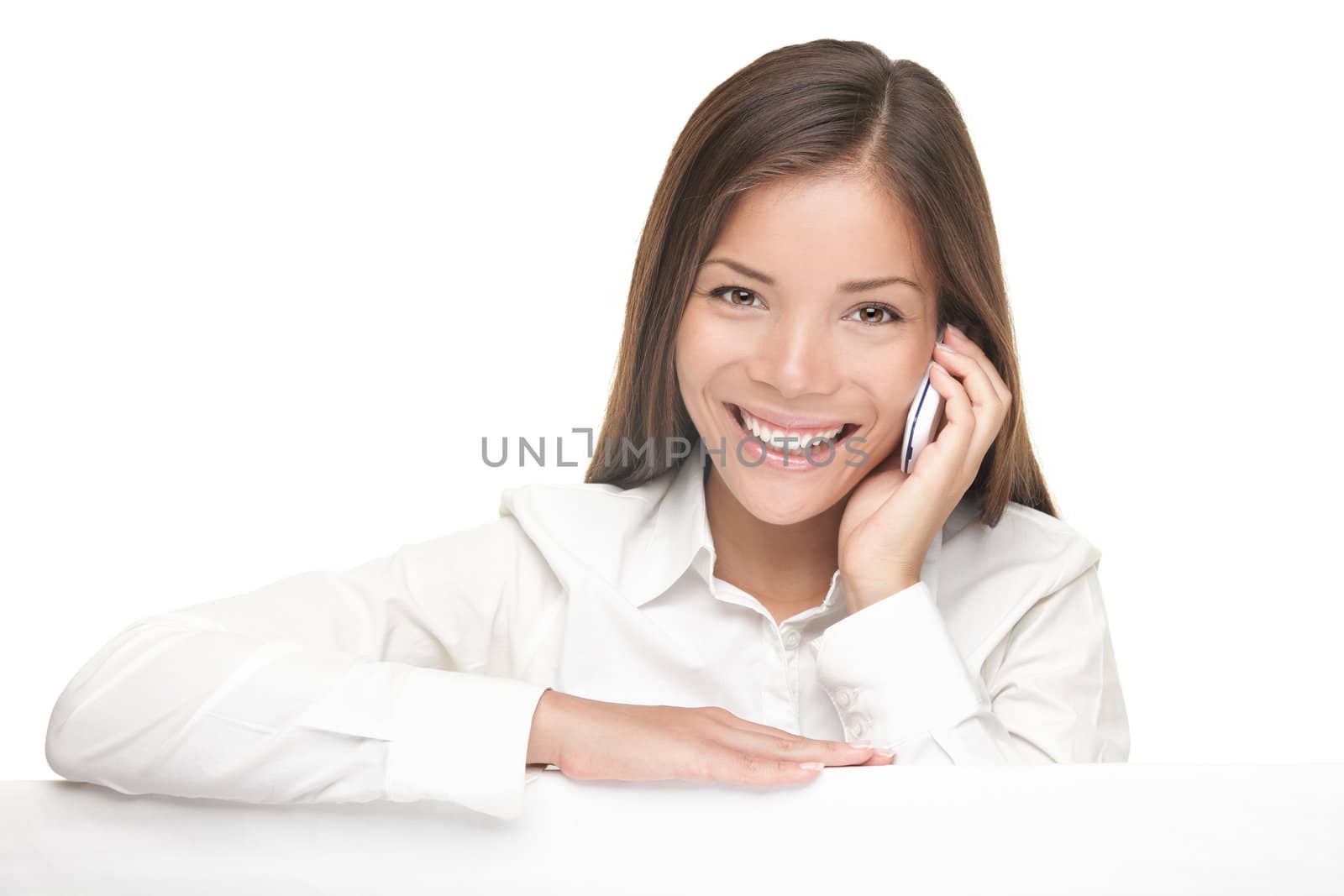 Woman showing billboard sign while talking on mobile phone and smiling. Young beautiful woman standing behind blank white billboard. Casual young Asian / Caucasian female model with very friendly relaxed smile. Isolated on white background 