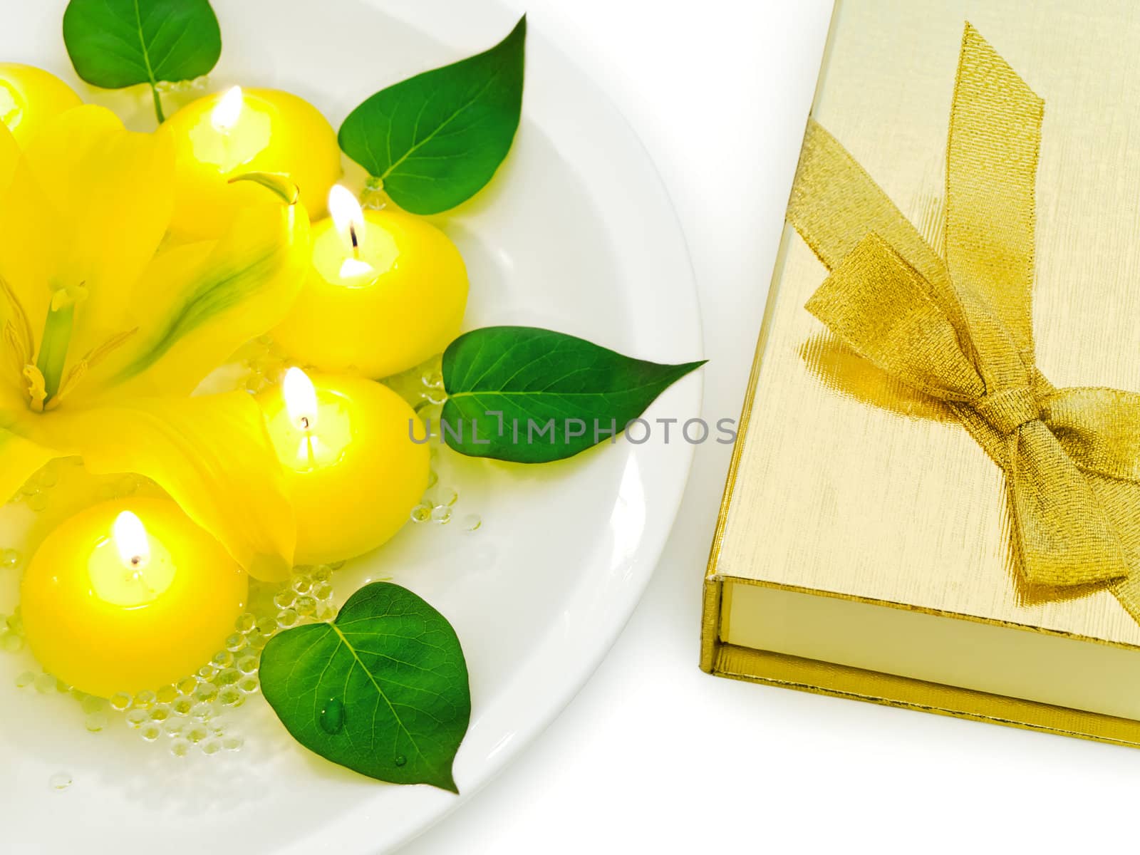 floral decoration with lily, candles and green leaves near golden gift box over white 