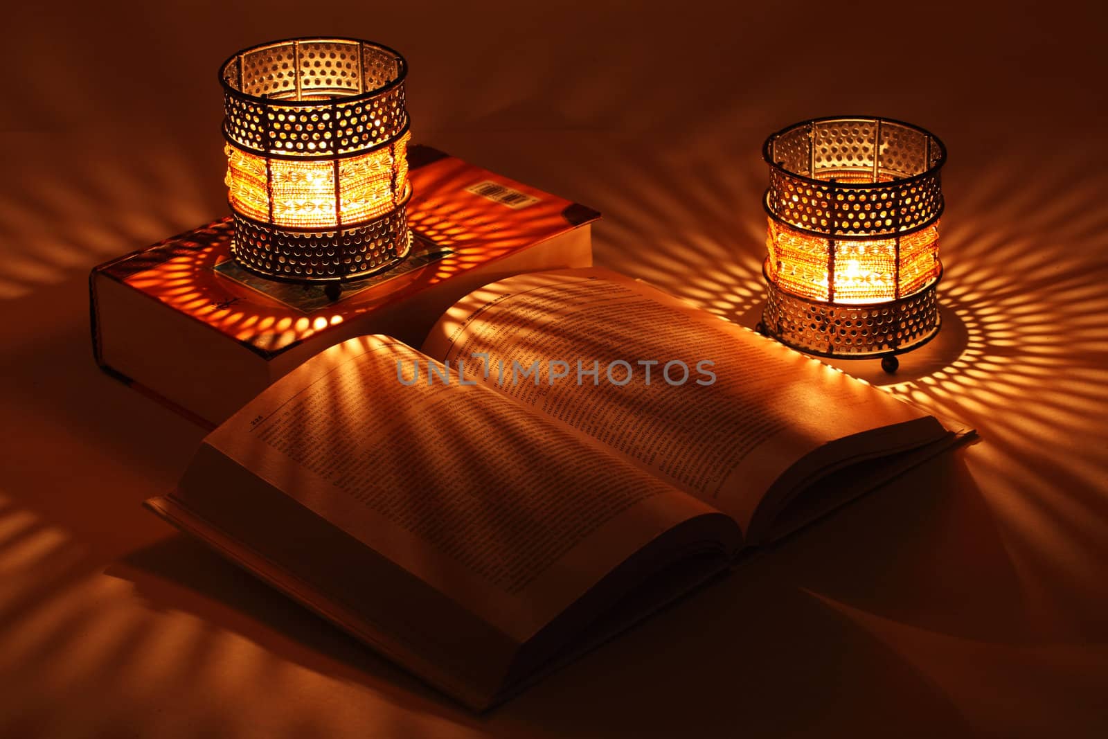 Two old-style candlesticks with flaming candle inside, one on big book and one open book in the dark