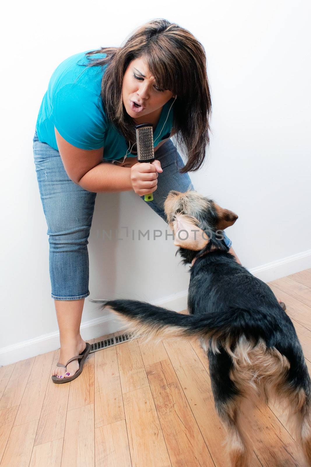A Hispanic woman singing on a pretend microphone that is actually a hair brush to the dog while listening to music playing through her stereo earbud headphones. 