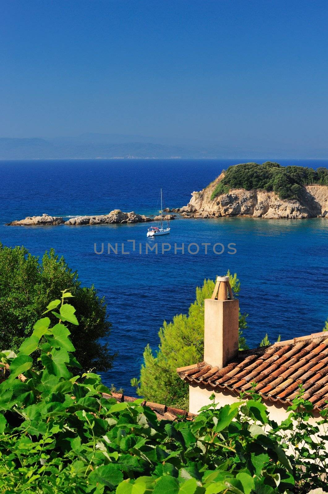 Morning view from a Greek island in the Aegean Sea, which includes part of roofed house, a small island, an anchored sailyacht. Room for text on top.