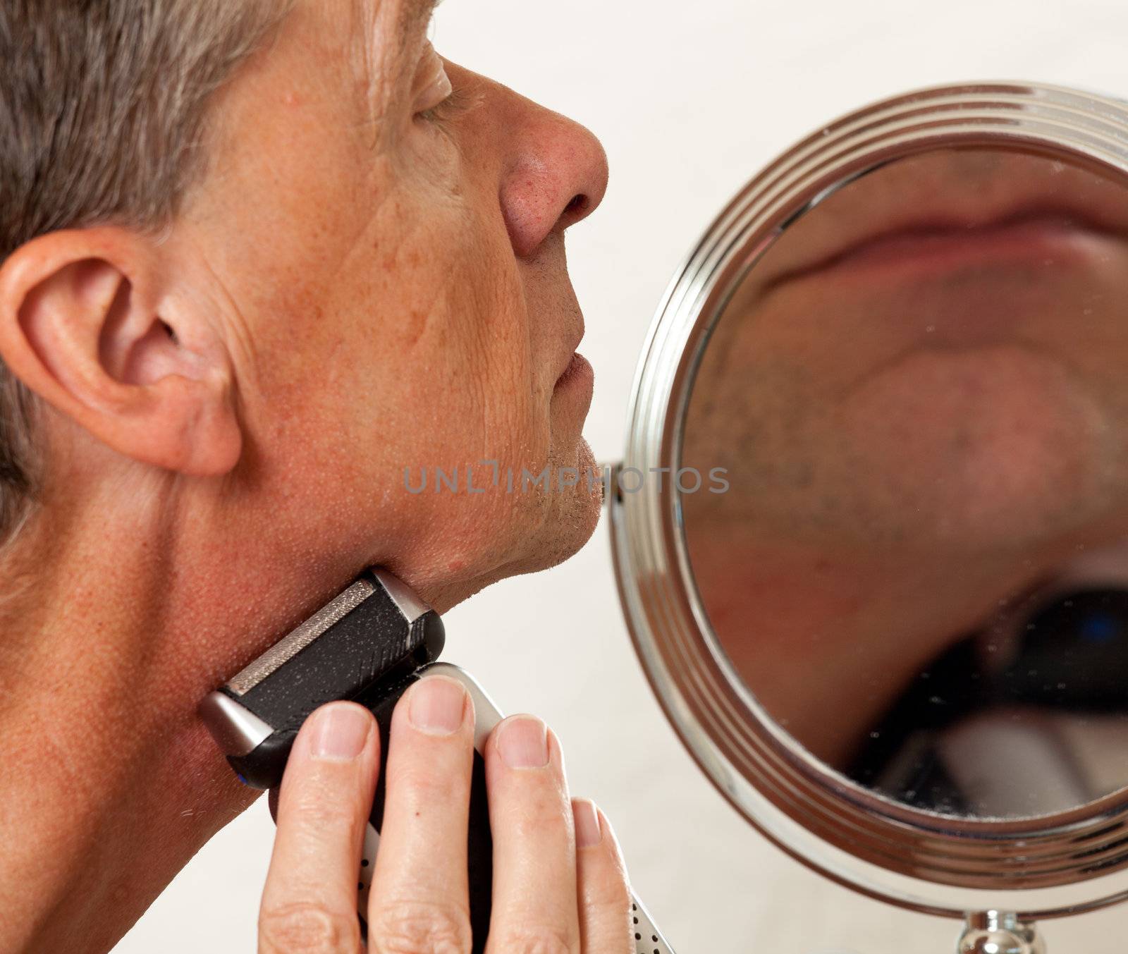 Retired male shaving with electic razor in front of magnifying mirror