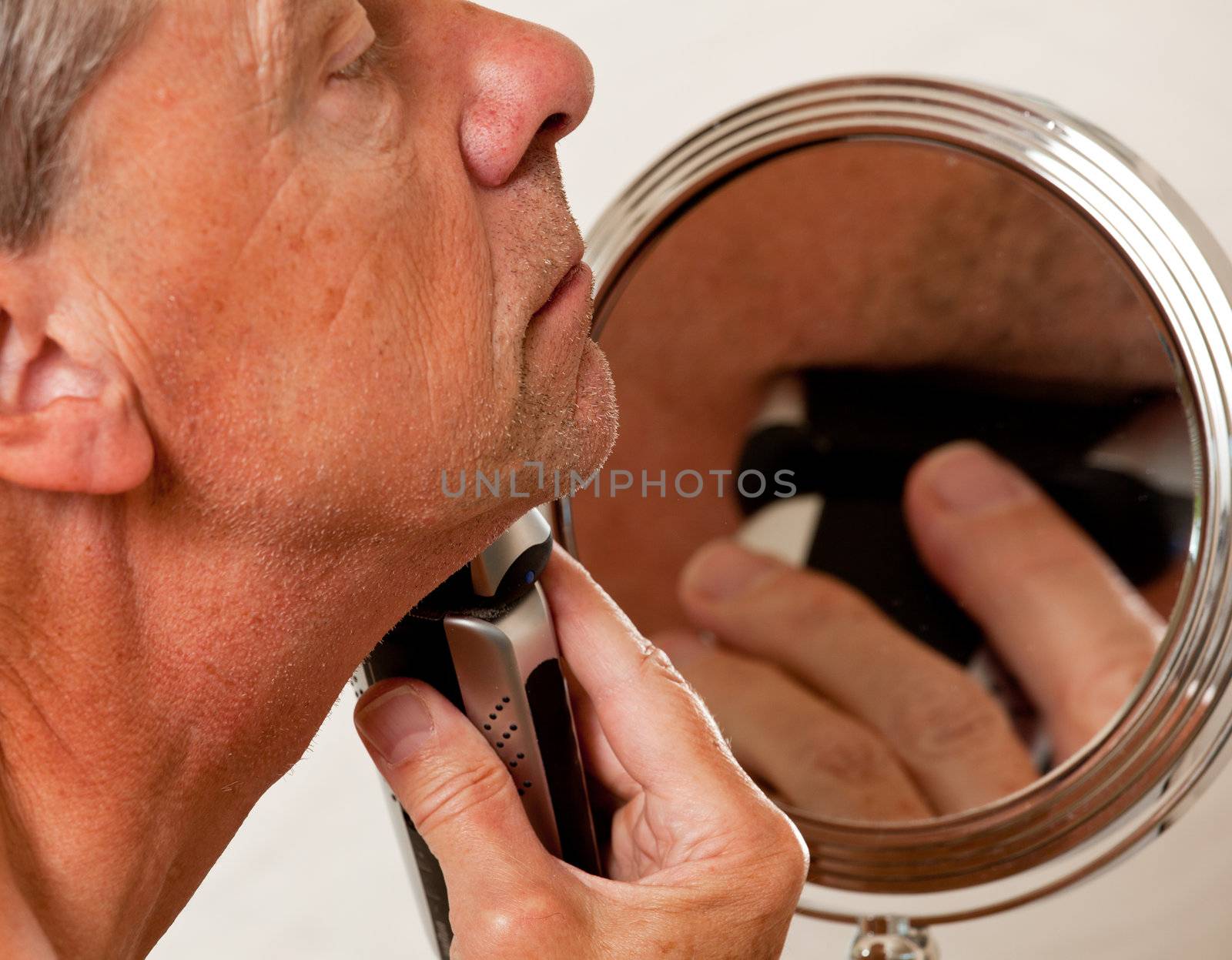 Retired male shaving with electic razor in front of magnifying mirror