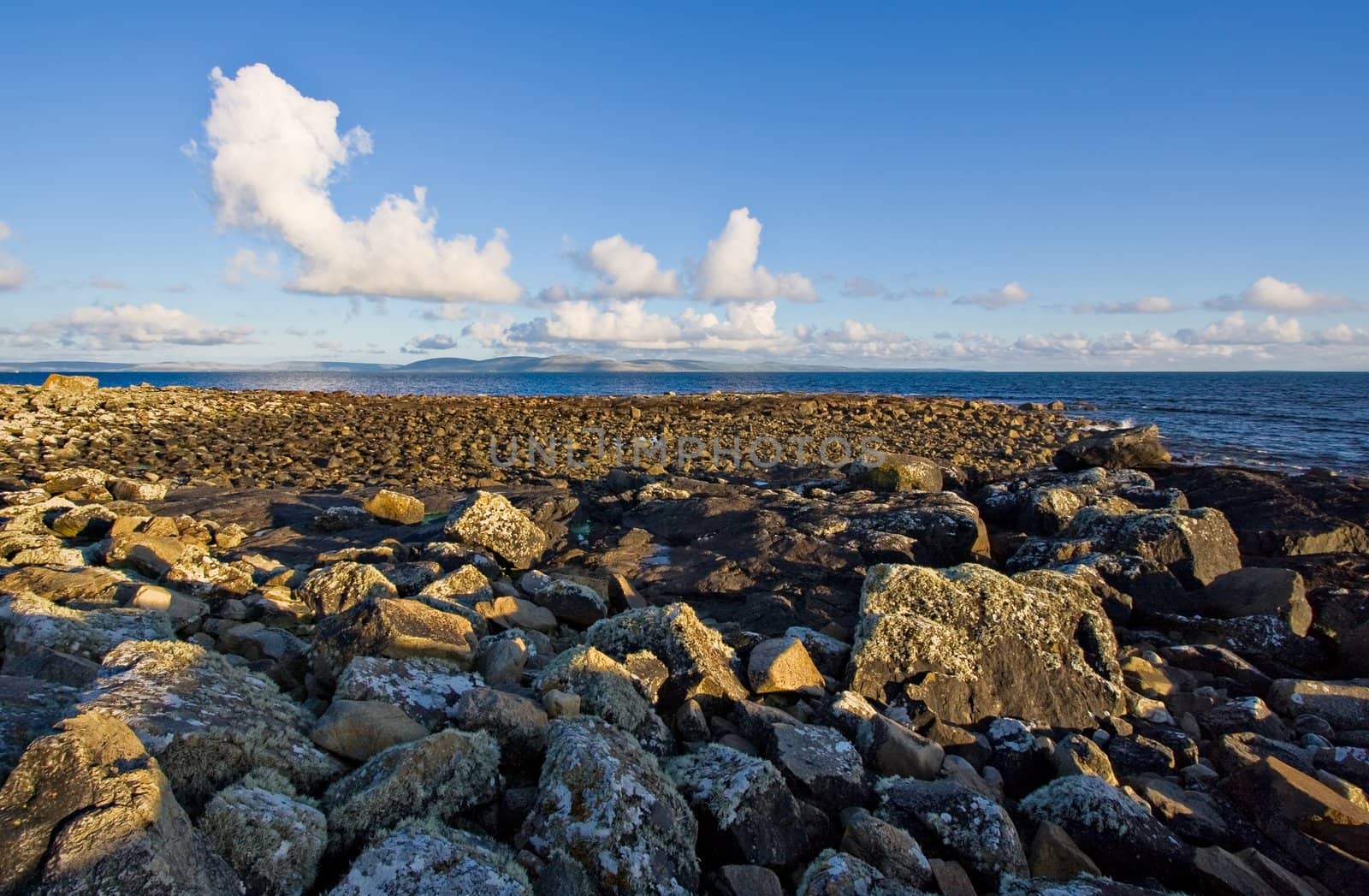 Galway Bay with a large rock jetty in the foreground. The Burren can be seen in the background with cumulus clouds against a blue sky.