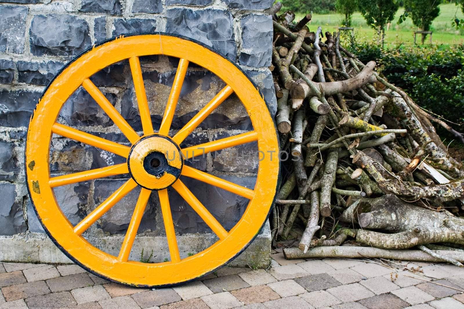 An old antique yellow wooden wagon wheel leaning against a stone wall. There is a pile of tree branches laying next to it.