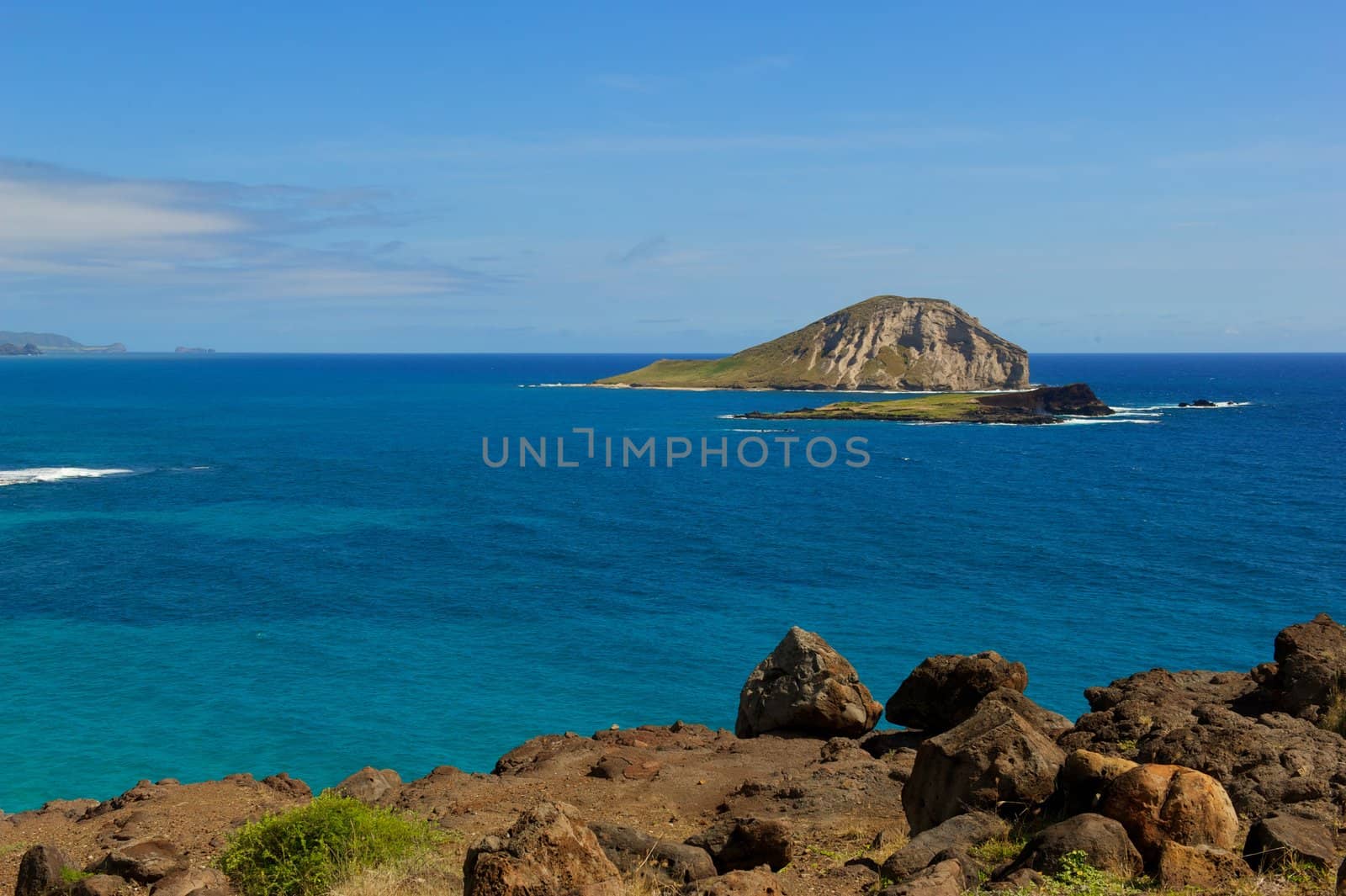 An island view with a beautiful bright, blue ocean, some grass patches, some rocks and sky.