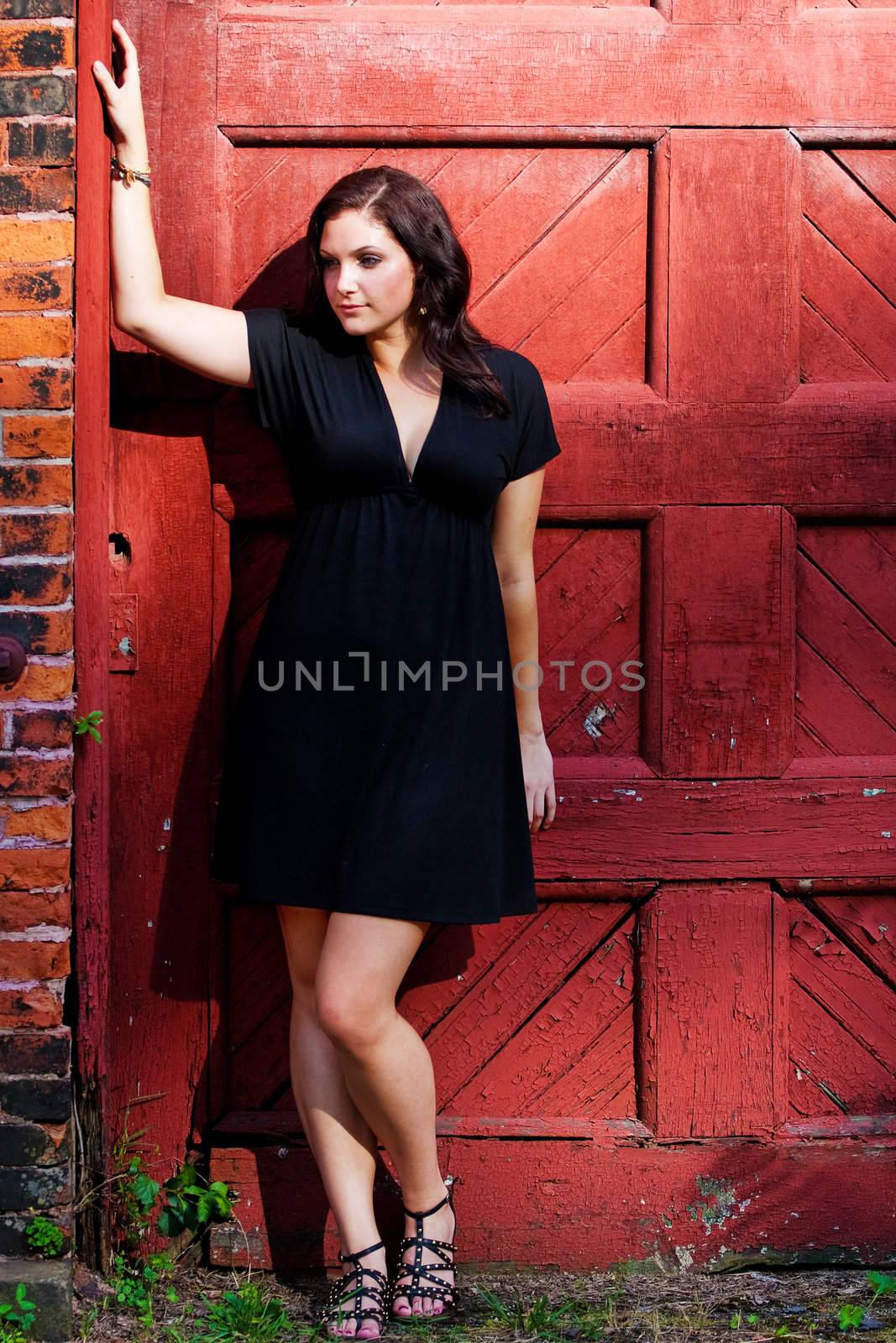 A pretty young woman in a black dress leaning against an old red doorway. 