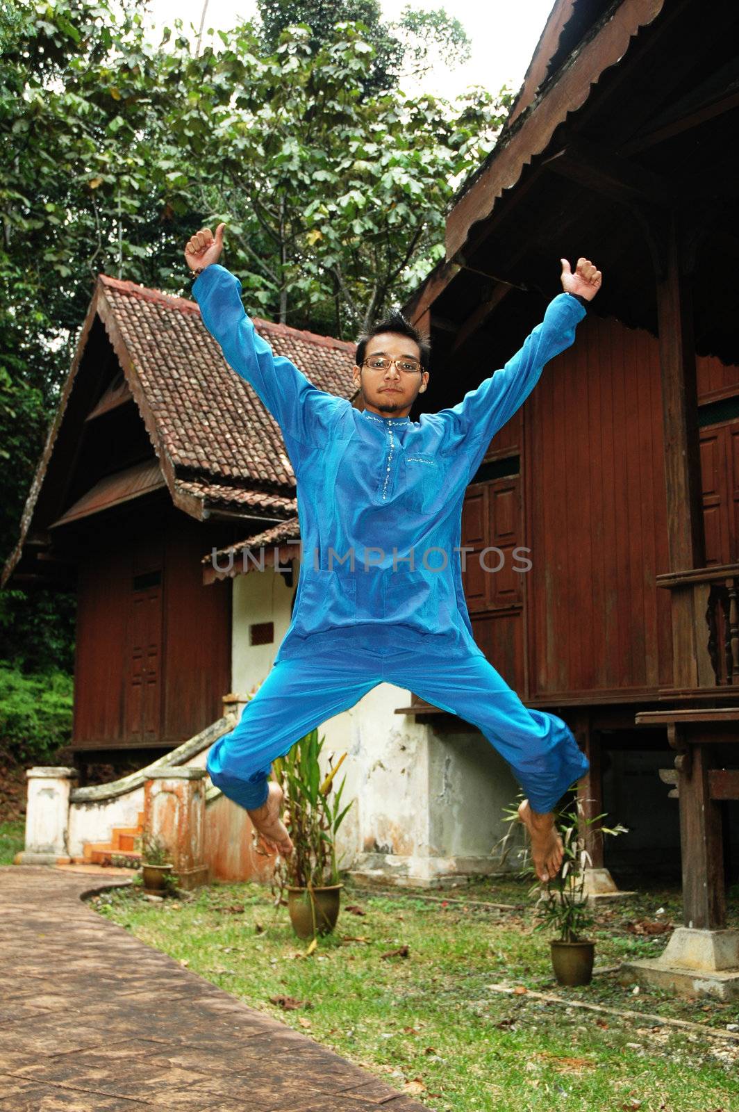 Asian male in traditional Baju Melayu jumping excitedly in front of a village house