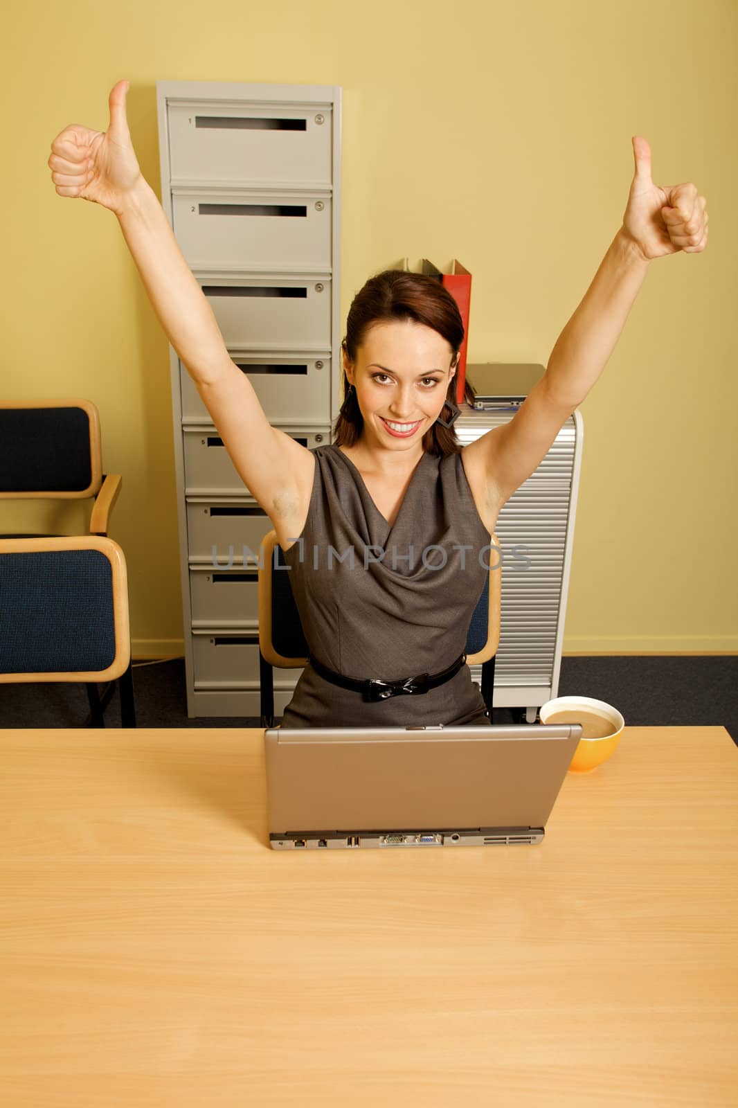 Portrait of businesswoman showing thumbs up sign, cup and laptop on desk