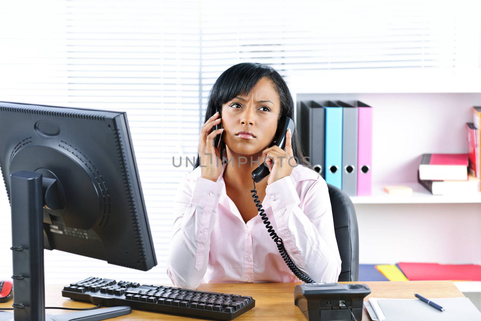 Black businesswoman using two phones at desk by elenathewise