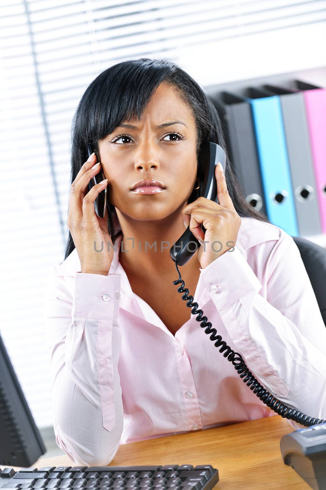 Black businesswoman using two phones at desk by elenathewise