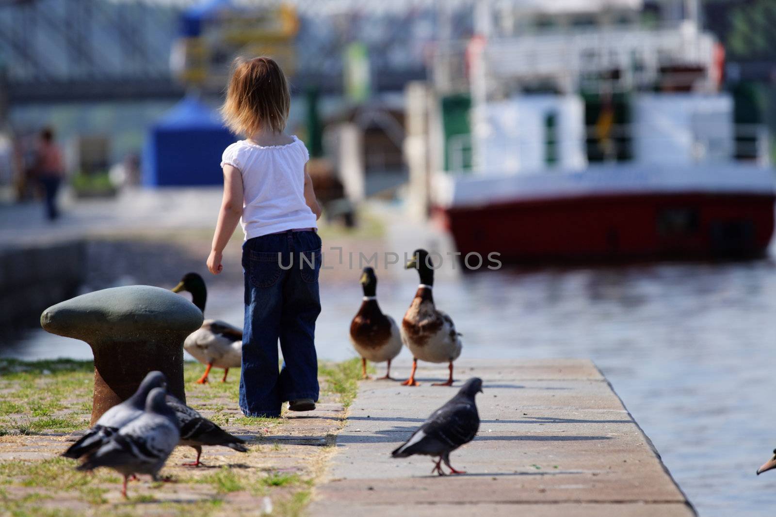 Little beauty girl feedind wild ducks at landing stage or river embankment at sunny day. We see ship not in fokus at background and doves. Prague, Vltava.