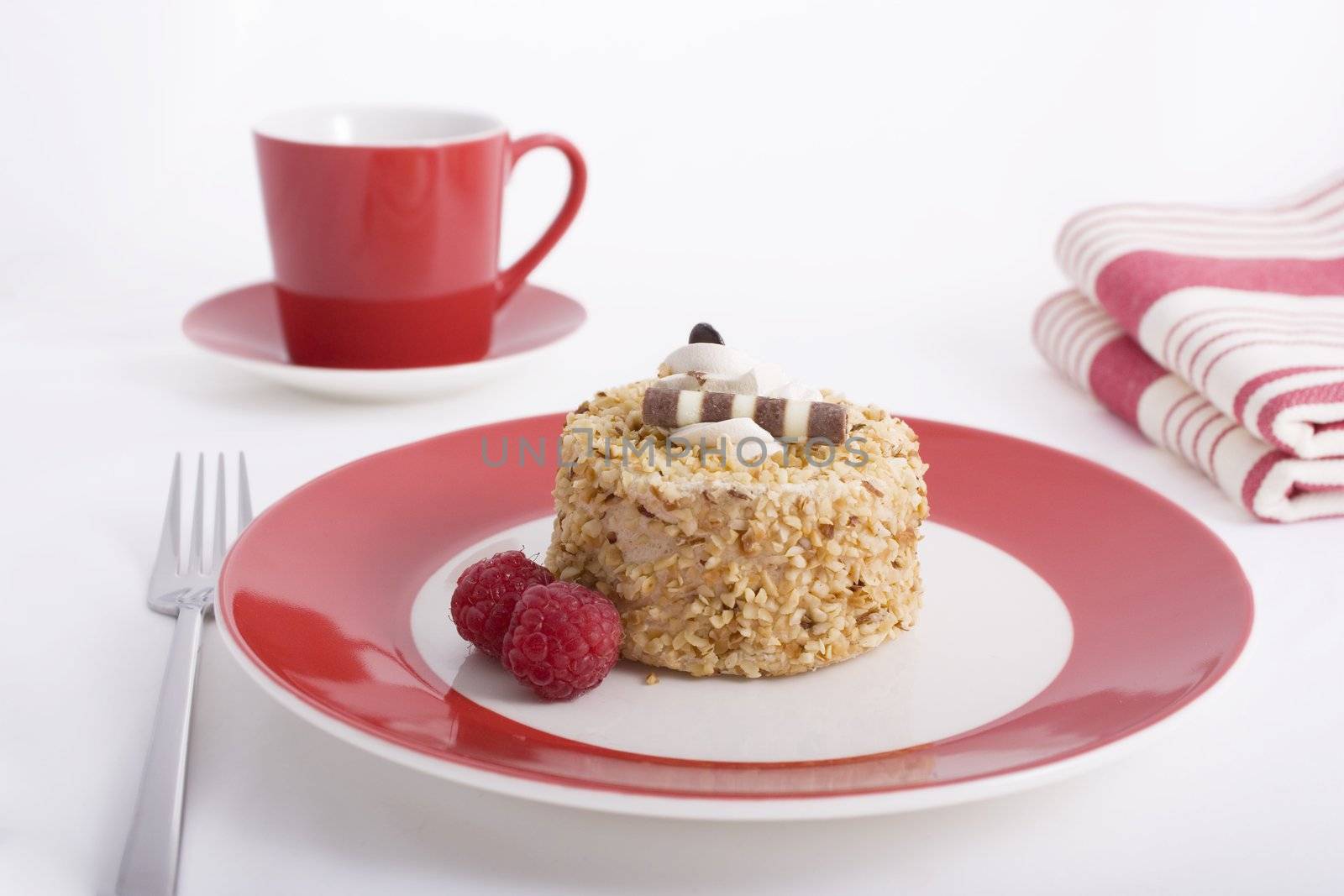 One serving of Hazelnut cake topped with cream and garnished with raspberries on a red plate.  A cup of coffee and napkins in the background.