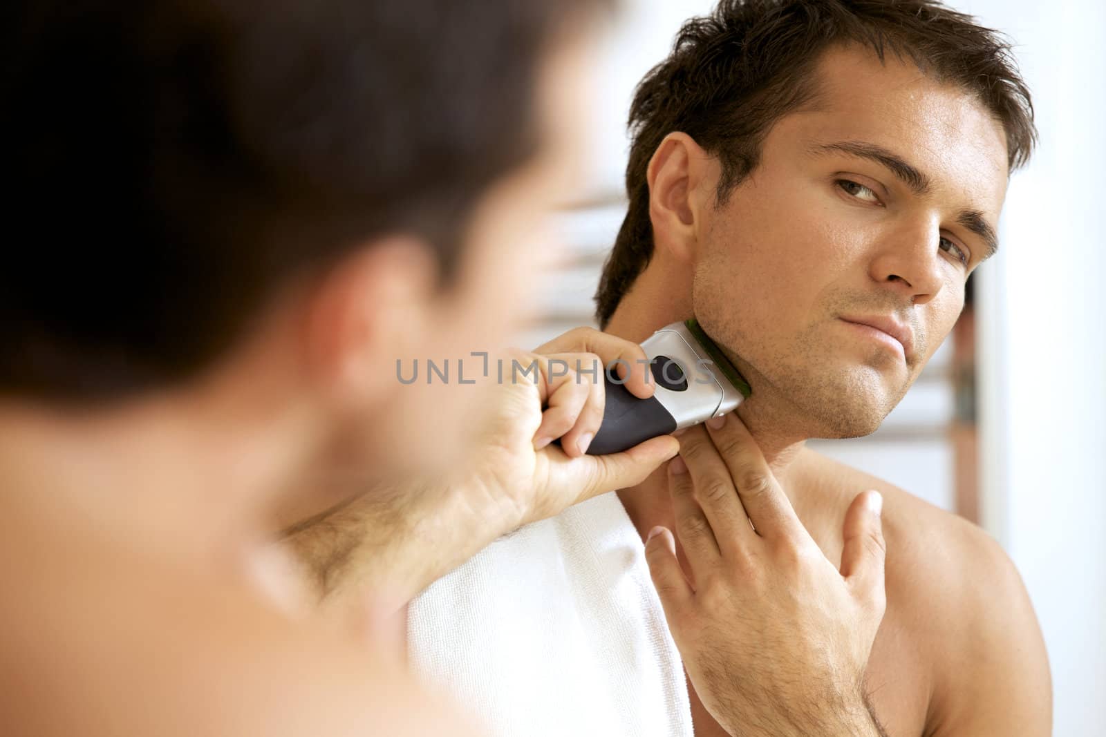 Reflection of young man in mirror shaving with electric shaver
