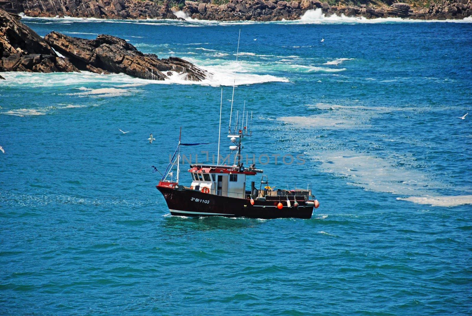 A boat travelling on the blue ocean, with many birds flying, and big rocs in the background.