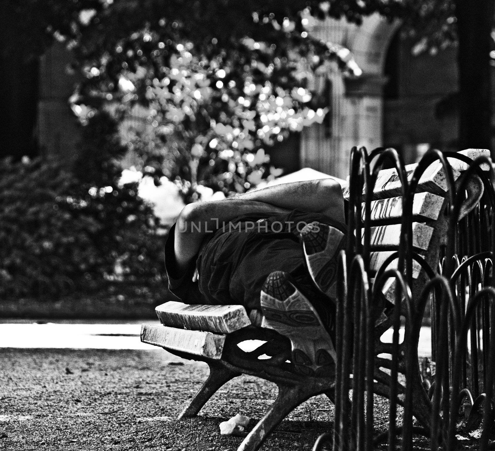 A man is sleeping on a bench, we can see his feet, legs and his crossed arms. The photograph is in black and white.
