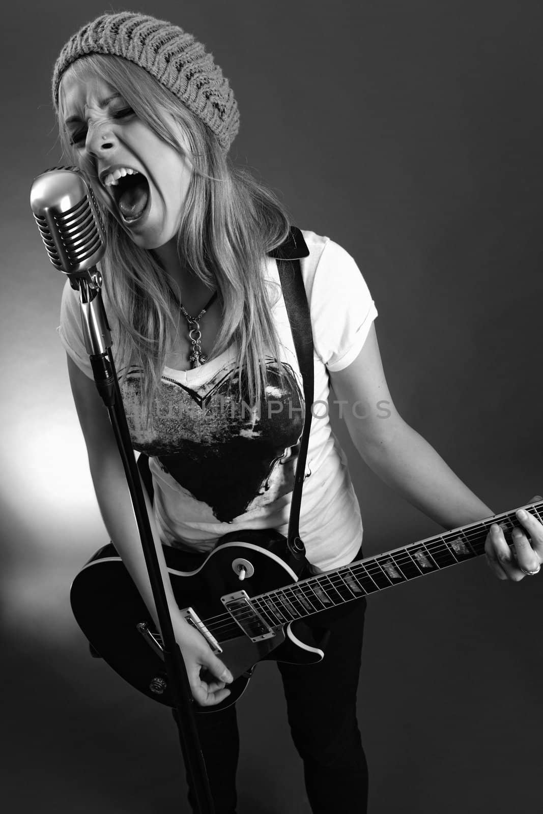 Black and white photo of a blond female screaming into an old microphone and playing electric guitar.  High contrast with film grain added.