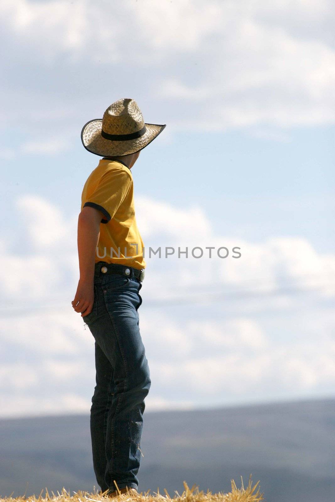 Male cowboy standing on straw stack looking away into the distance with yellow shirt, blue jeans, and hat