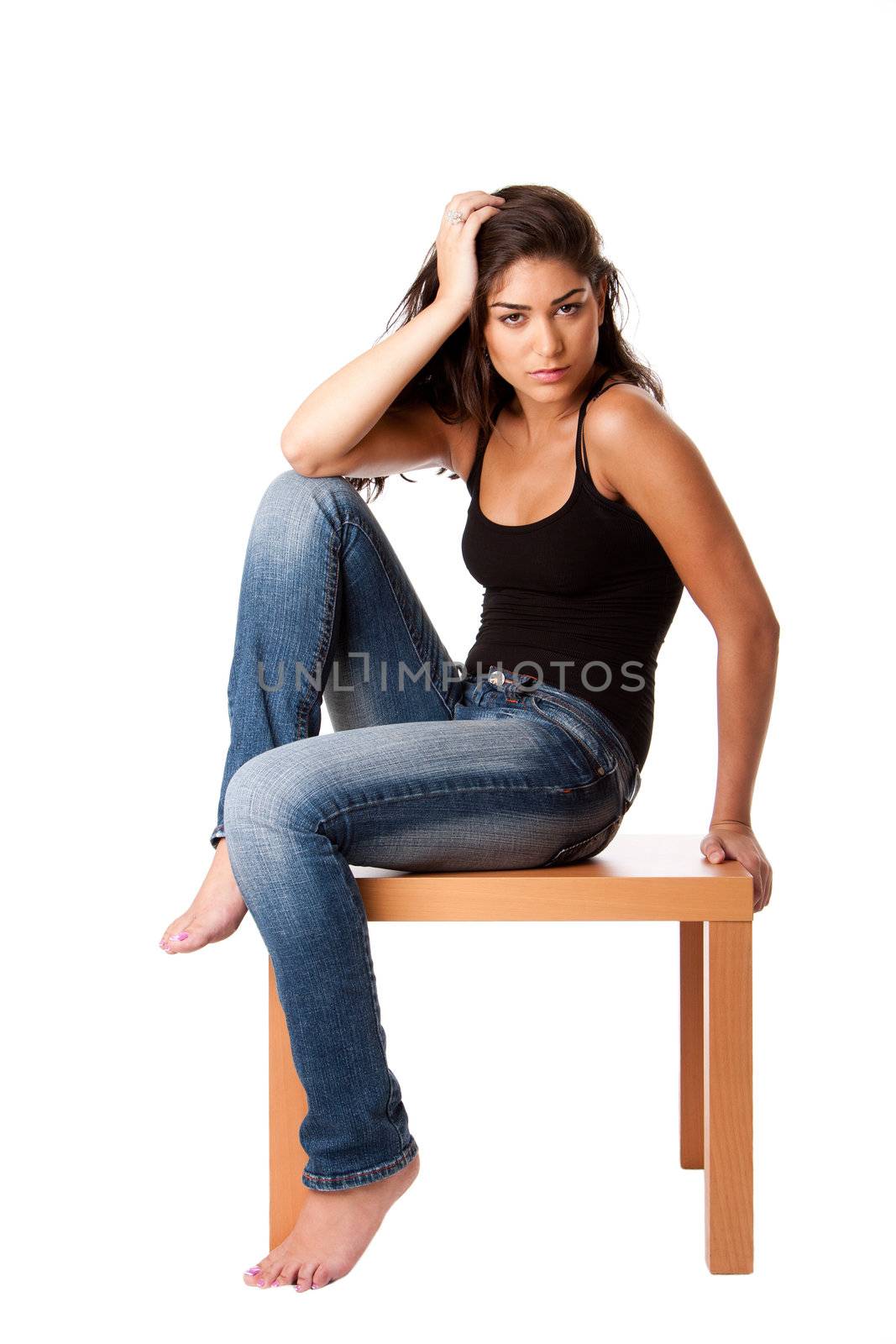 Beautiful fashion woman sitting on wooden table wearing blue denim jeans and black tank top, isolated.