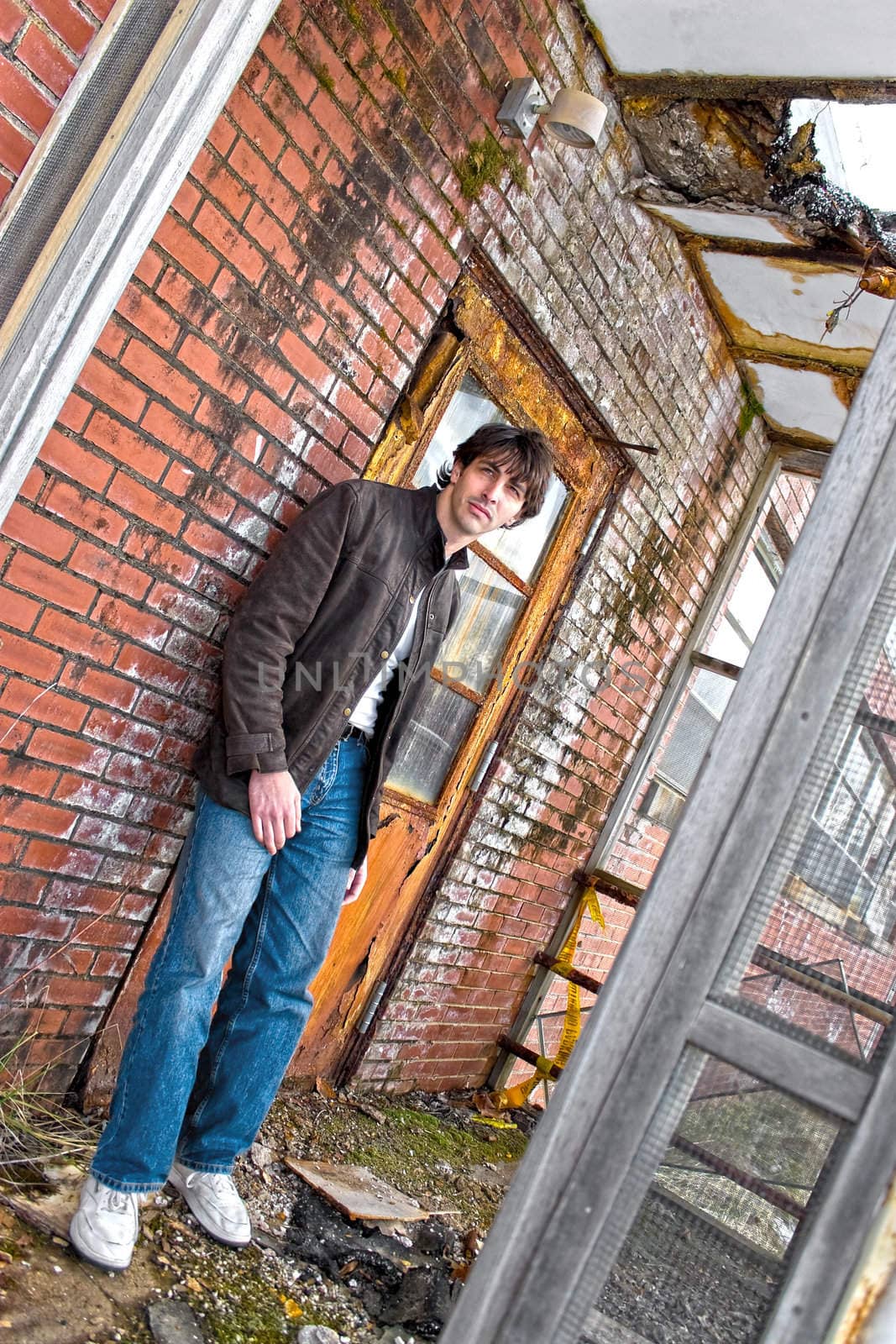 A young brunette man posing outside a worn and abandoned building.