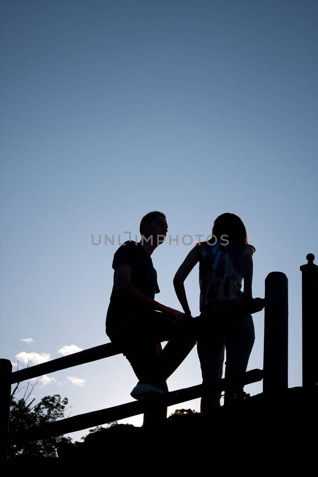 Silhouette of a young couple hanging out together outdoors by an old country fence during the early evening hours.