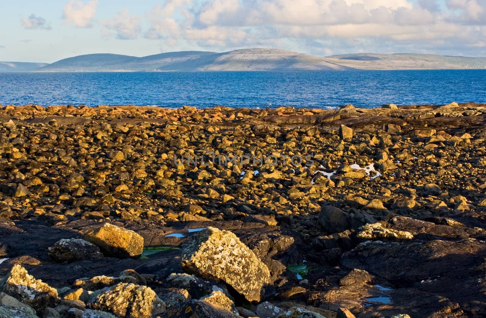Galway Bay in Ireland with The Burren across the bay. Photo is layered from front to back with Rocks, Galway Bay, The Burren, and sky.