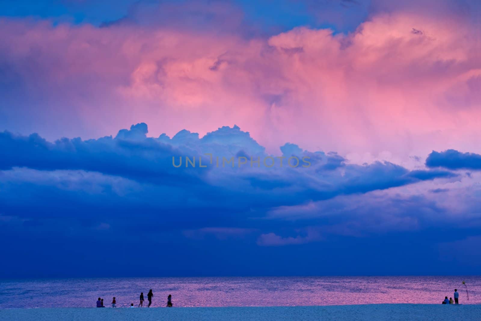 A beach scene in the evening at sunset. There are people on the beach with a dramatic stormy sky overhead. The last rays of sunlight are lighting some of the clouds.