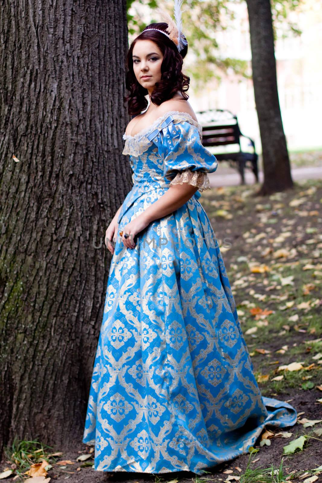 A portrait of lady in a blue baroque dress standing near a bole
