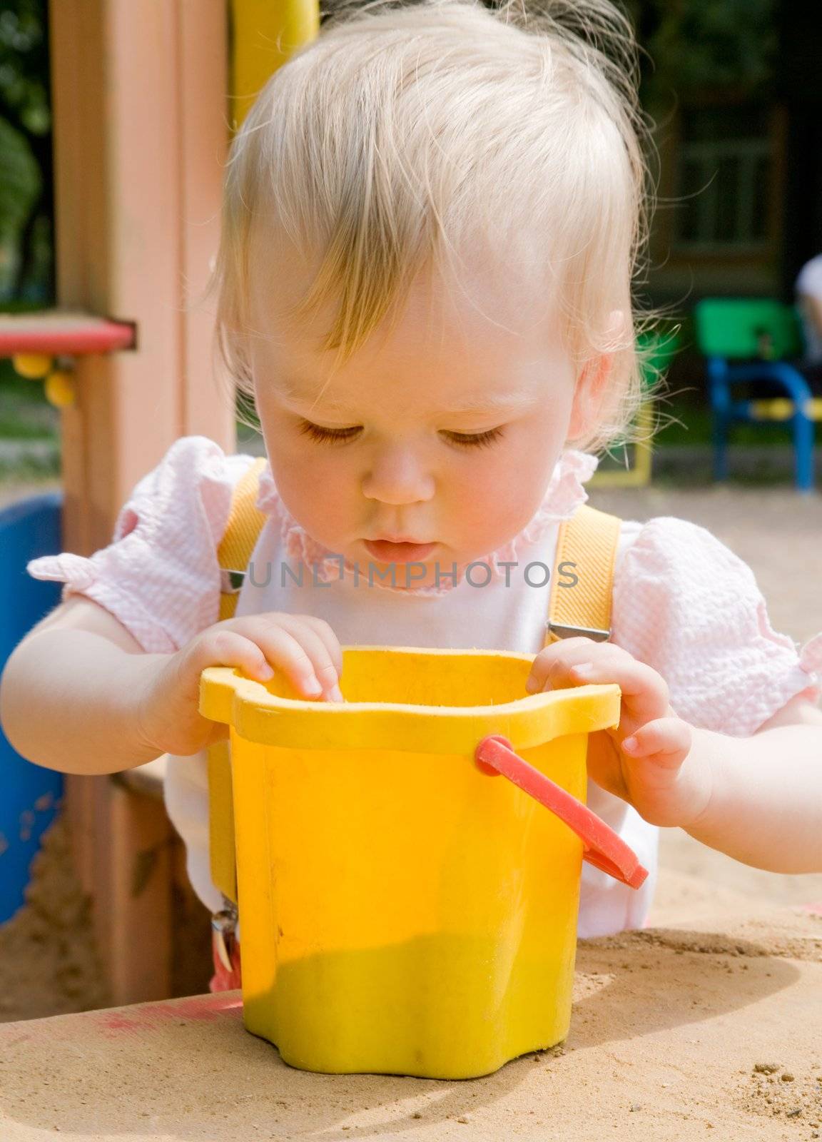 little girl looks in a yellow bucket with sand in a sandbox on a playground