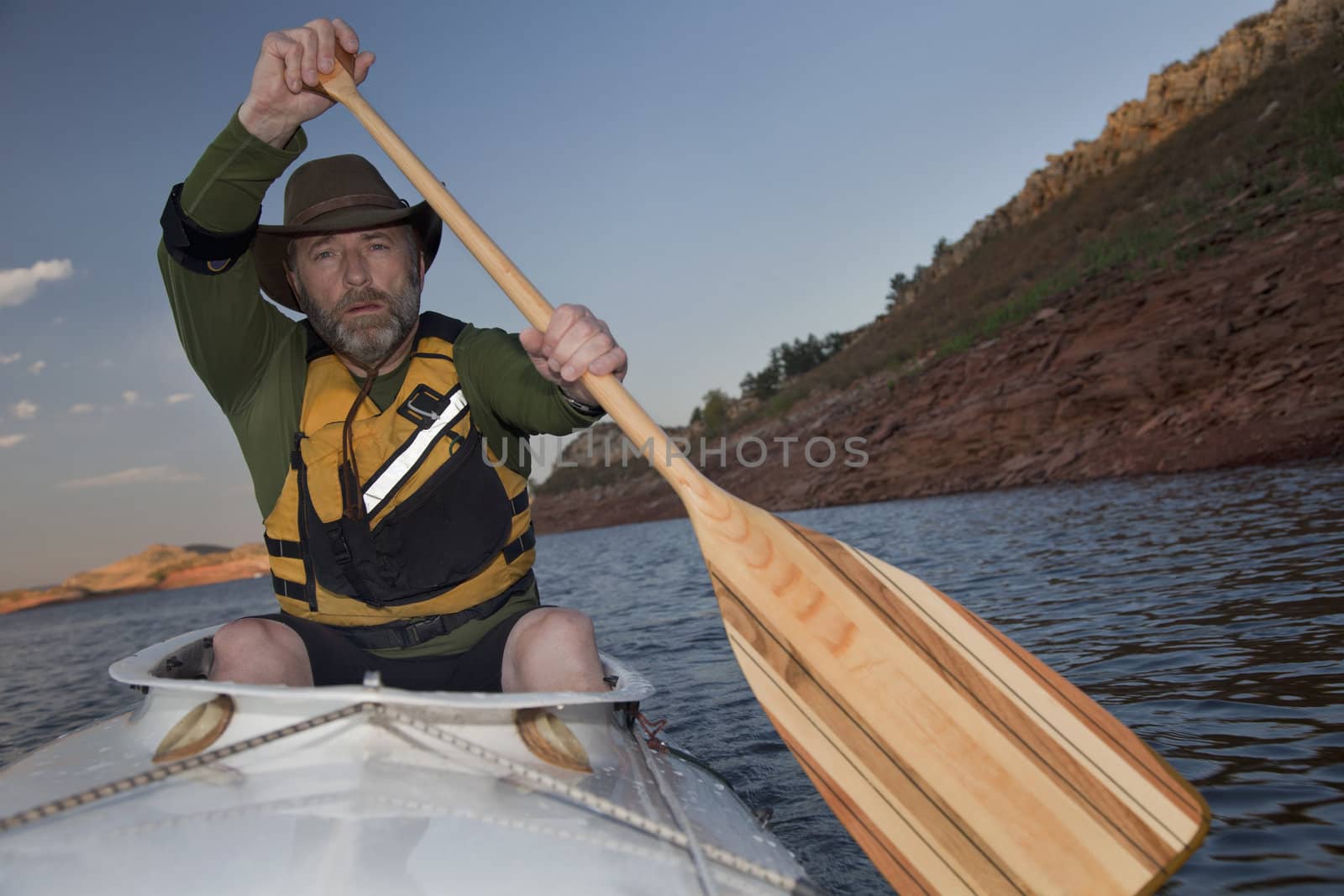 mature male paddling a white decked expedition canoe with wooden paddle on mountain lake with red sandstone cliffs (Horsetooth Reservoir near Fort Collins, Colorado)